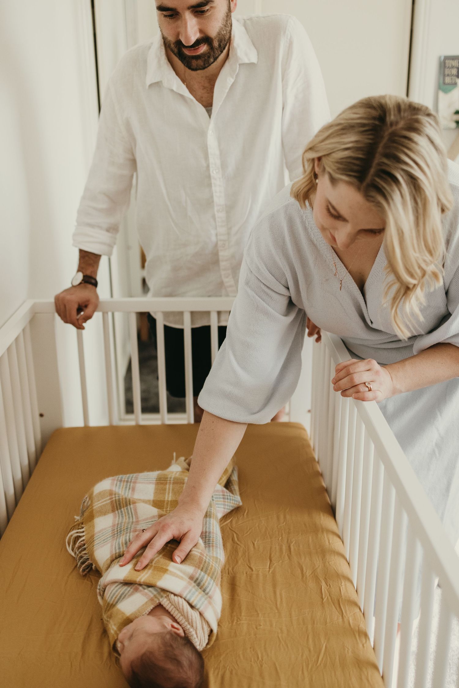 Parents stand over white crib with mustard sheets tending to their swaddled newborn.