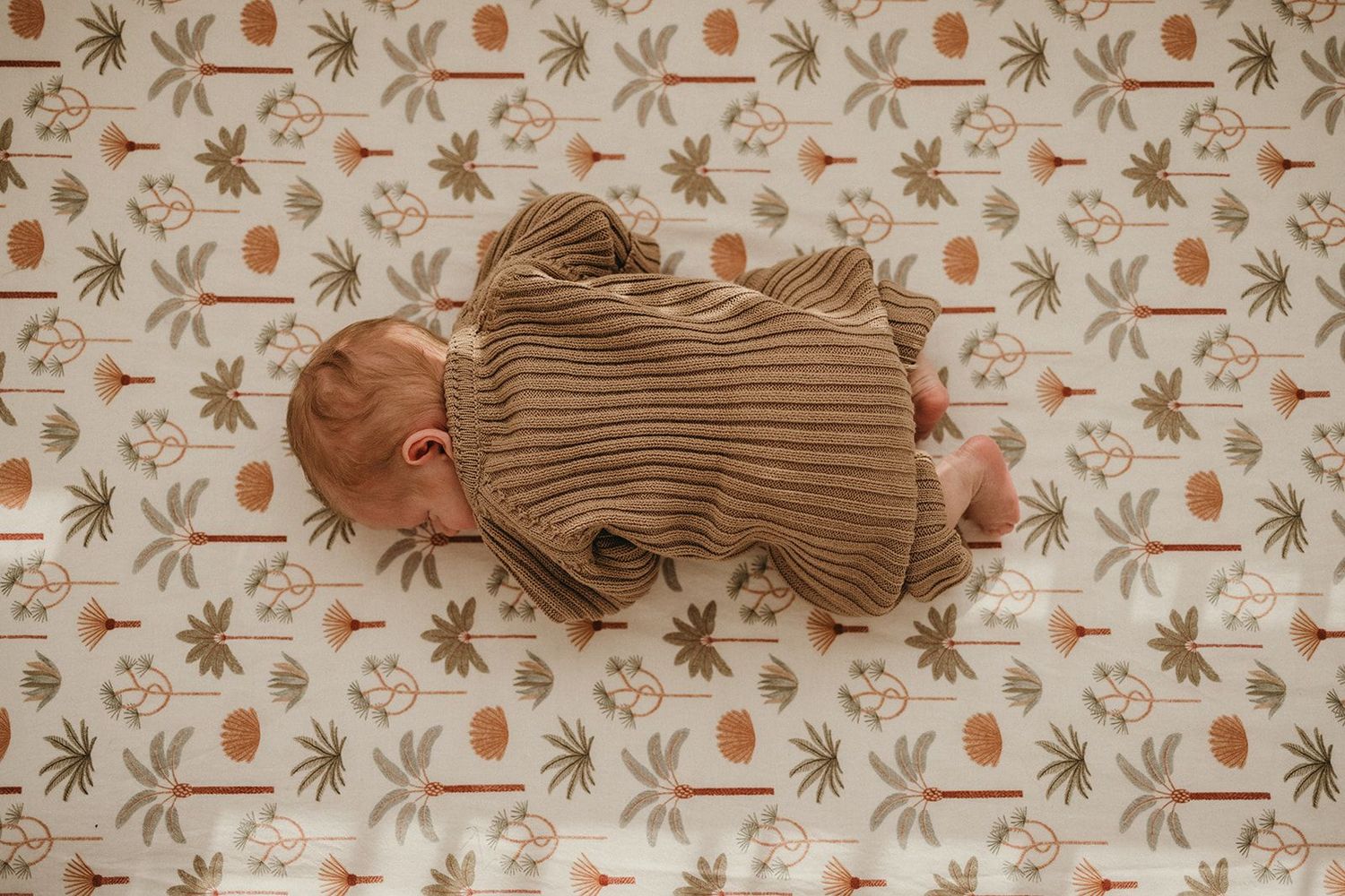 Newborn baby sleeping peacefully on a floral patterned blanket wearing a brown knit outfit, photographed from above.
