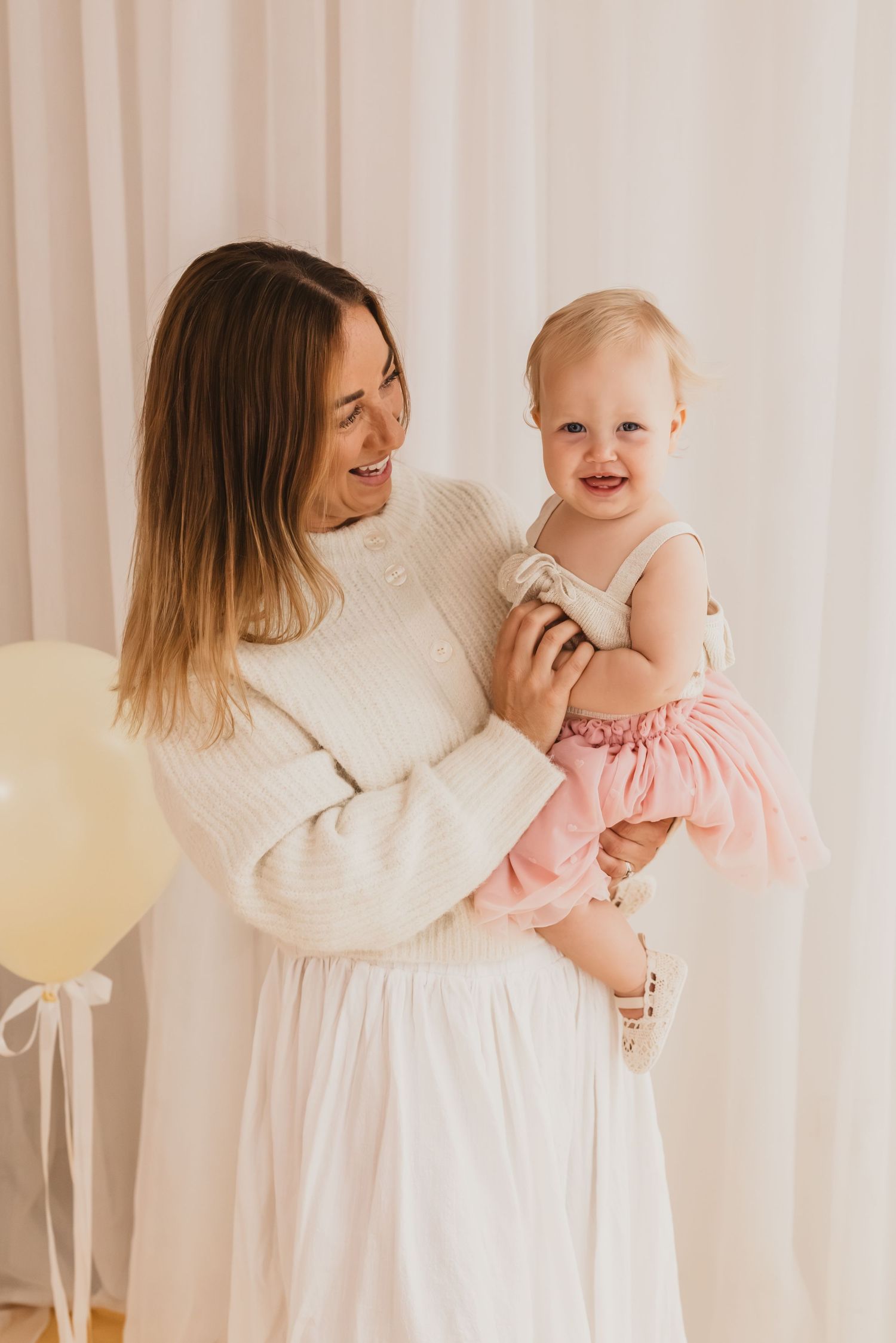 A smiling mother in a white dress holds her happy baby girl in a pink tutu skirt near pastel balloons.