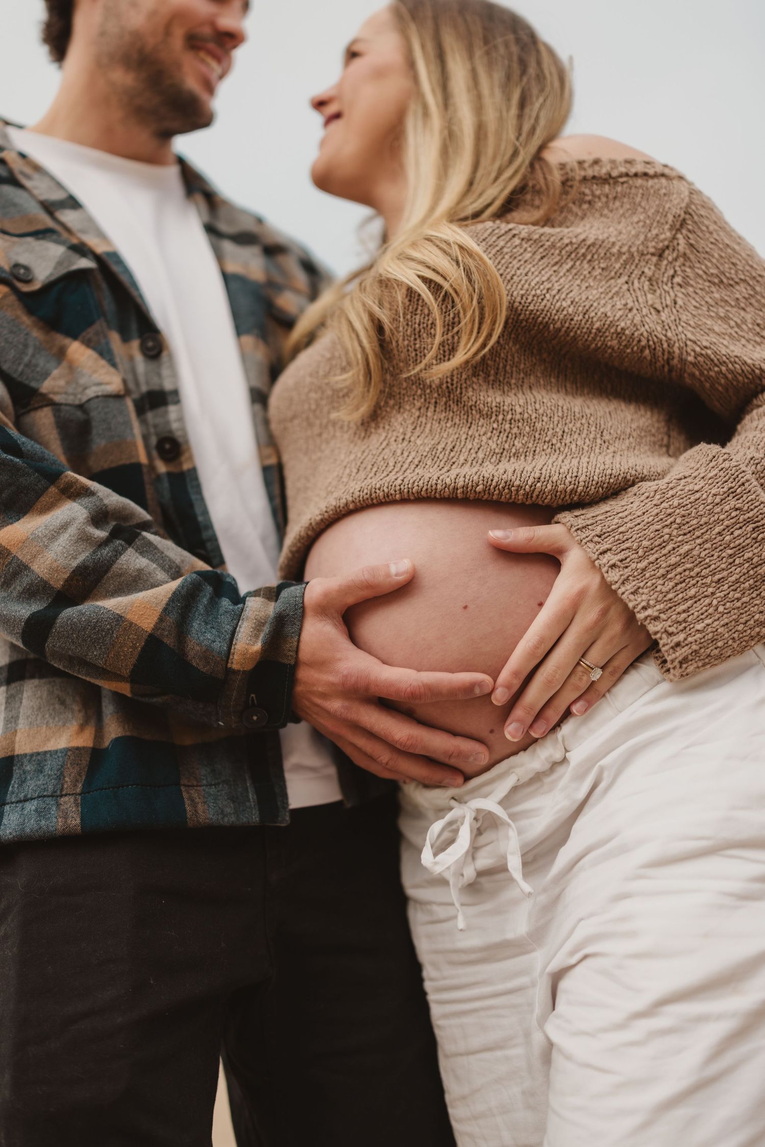 Couple tenderly cradling pregnant belly outdoors, wearing cozy flannel and knit sweater, sharing a joyful smile.