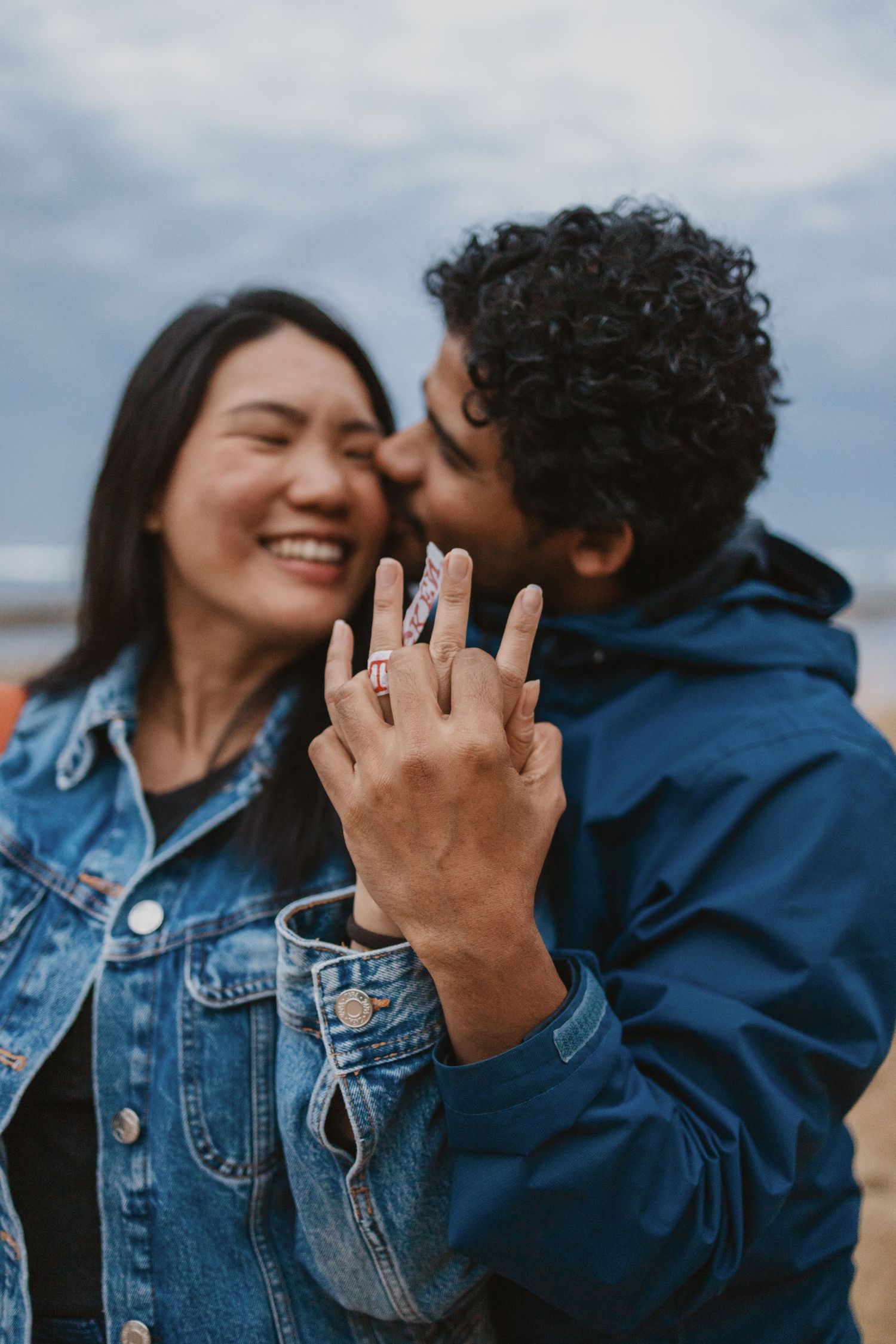 Joyful couple celebrating engagement on beach, woman showing diamond ring while man kisses her cheek.