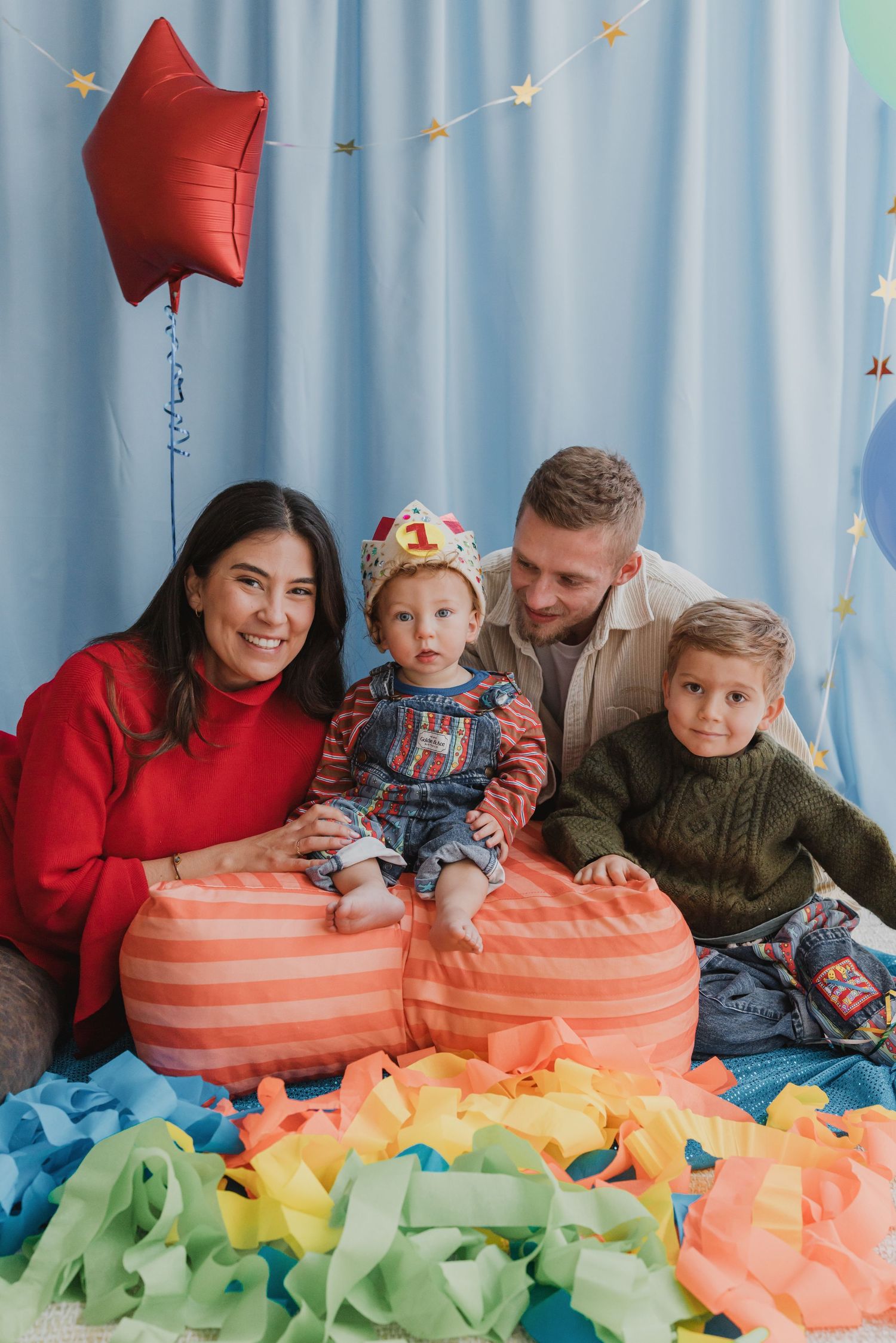 Family celebrating baby's first birthday with colorful confetti, balloons, and a crown in a festive blue backdrop.