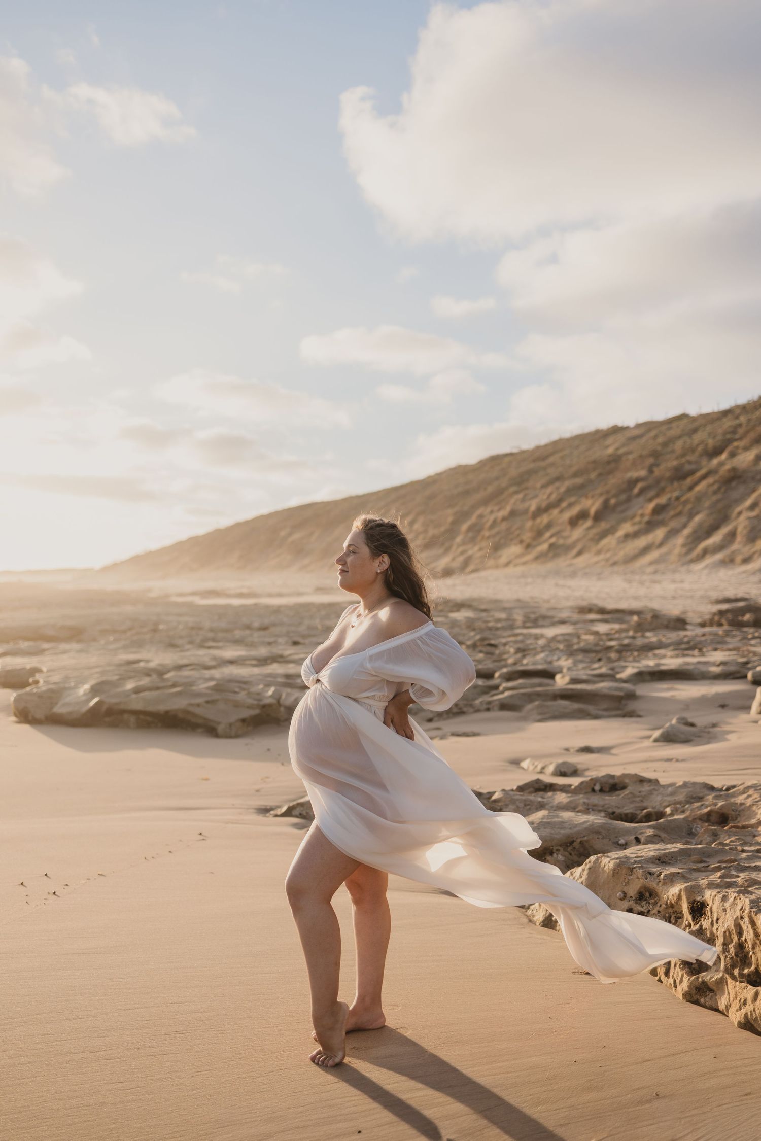 Pregnant woman in flowing white dress on sandy beach with rocky cliffs, golden hour light.