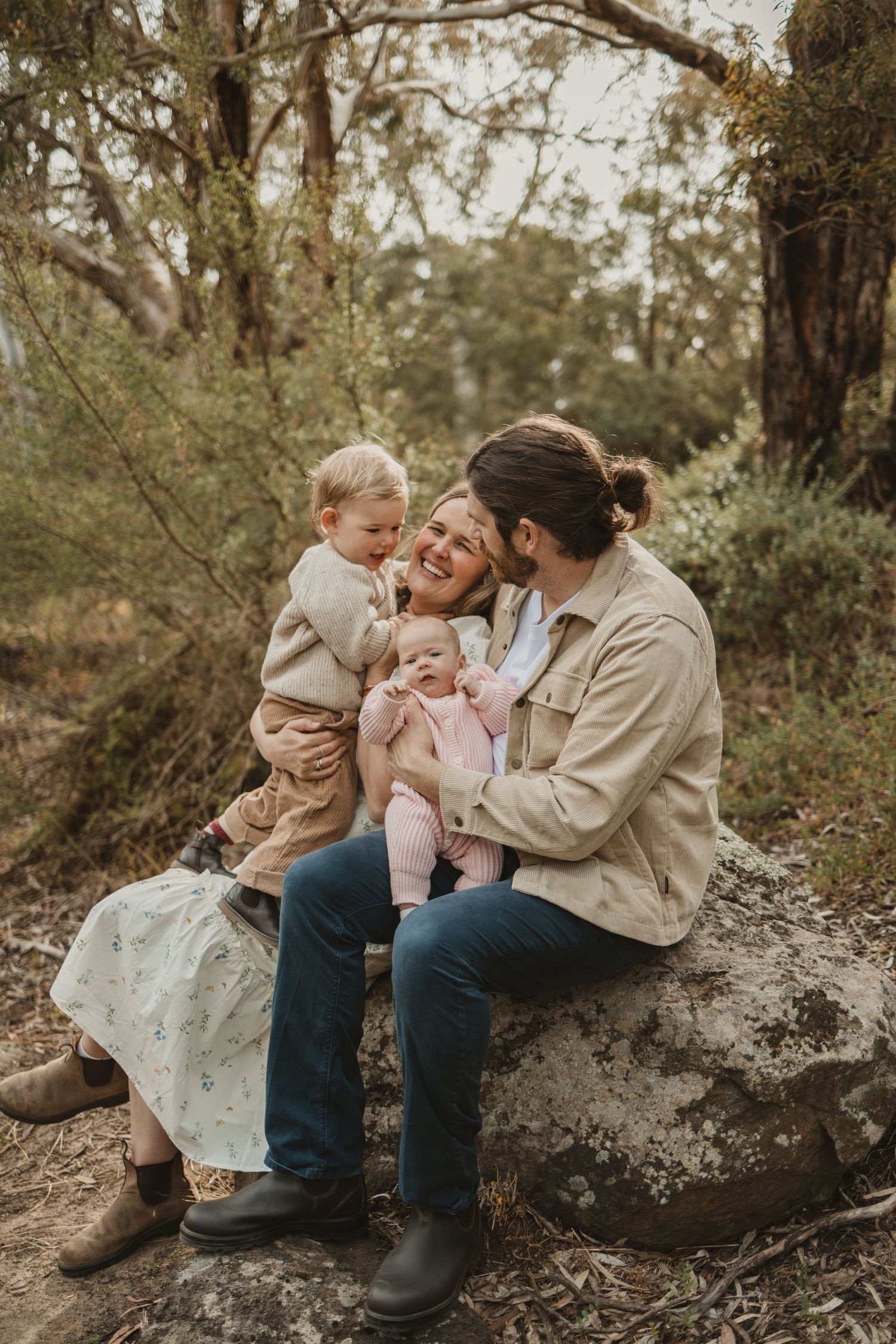 Happy family of four sitting on a rock in a forest, parents smiling with toddler and baby in nature.