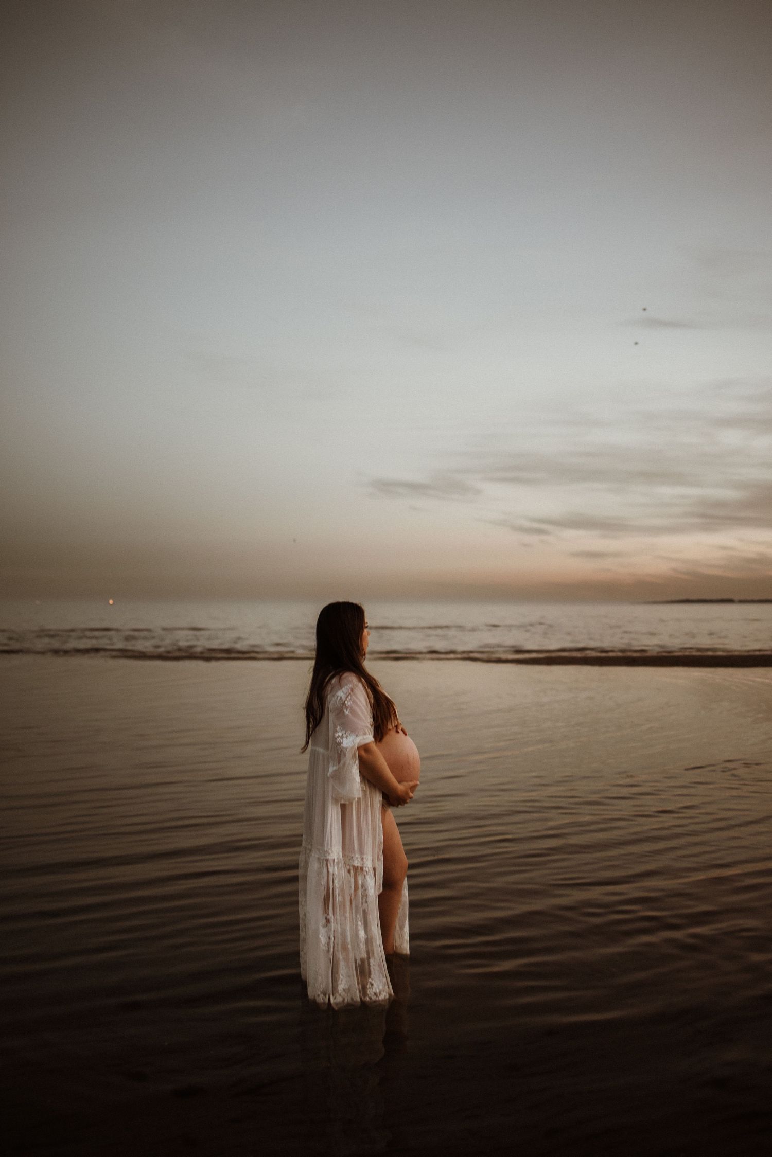 Pregnant woman in white lace dress standing in shallow ocean water at sunset, cradling her baby bump.