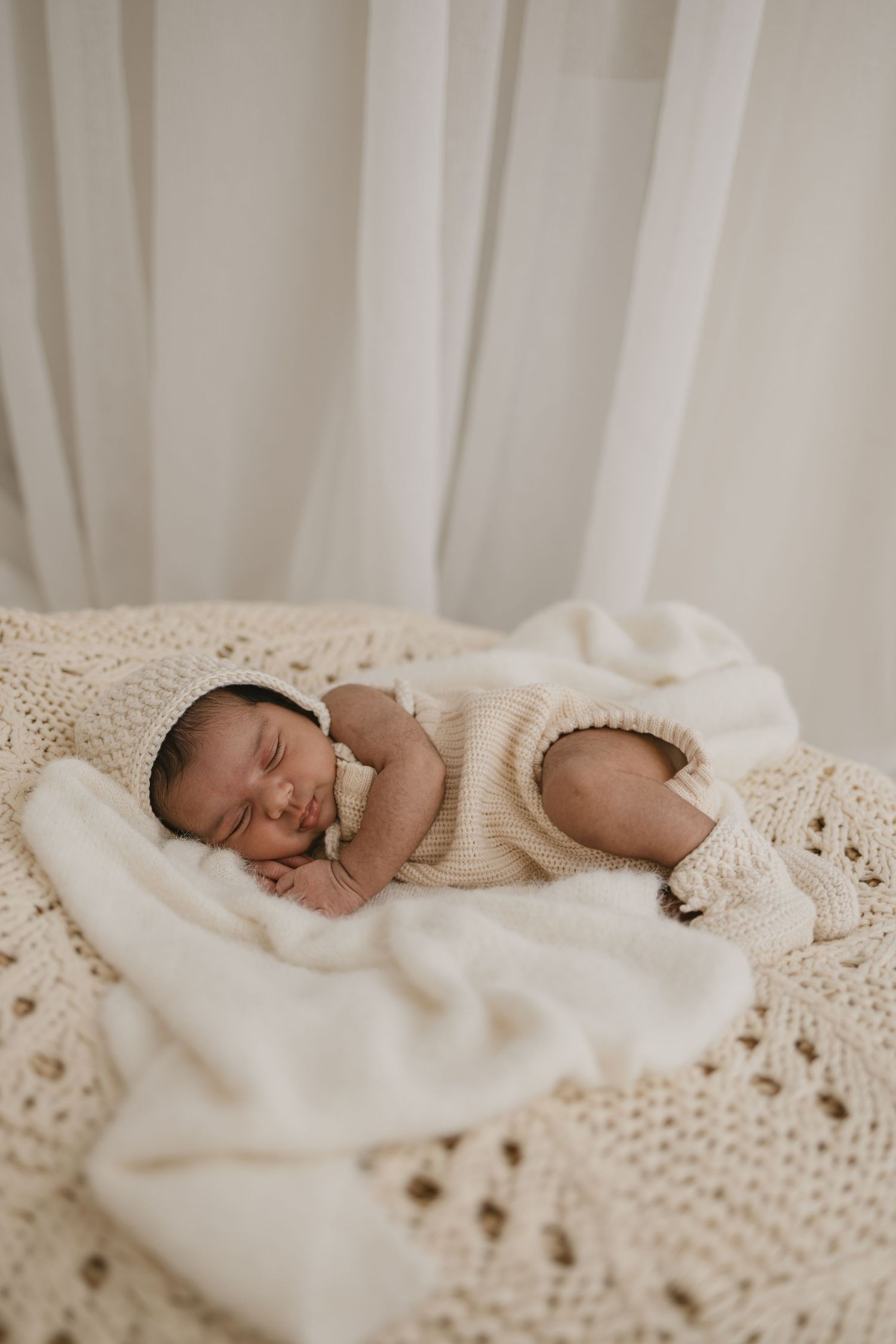Sleeping newborn baby in cream knit outfit resting on white crochet blanket with soft curtain backdrop.