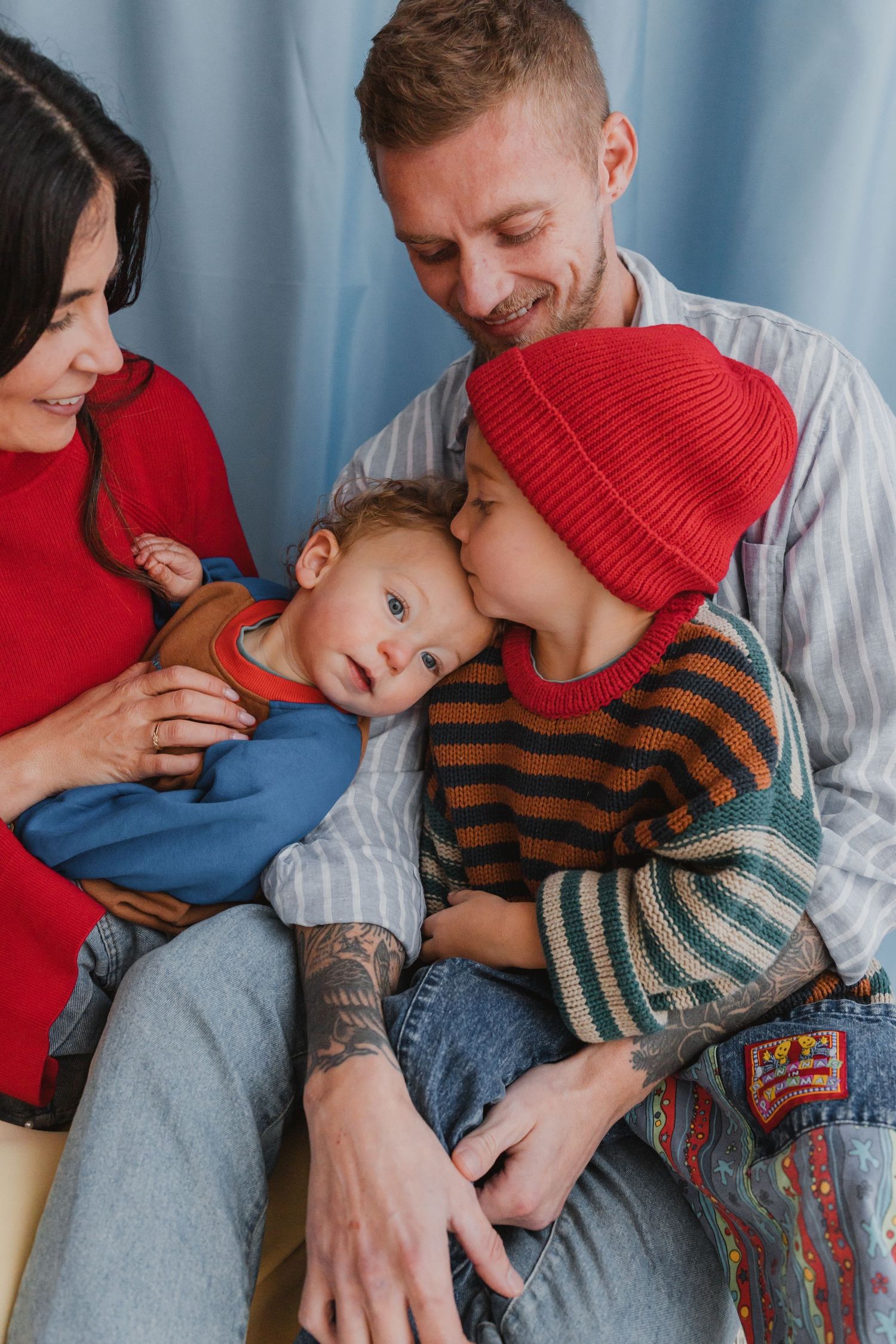 A happy family of four with tattooed dad, mom in red, older child in striped sweater kissing a baby.