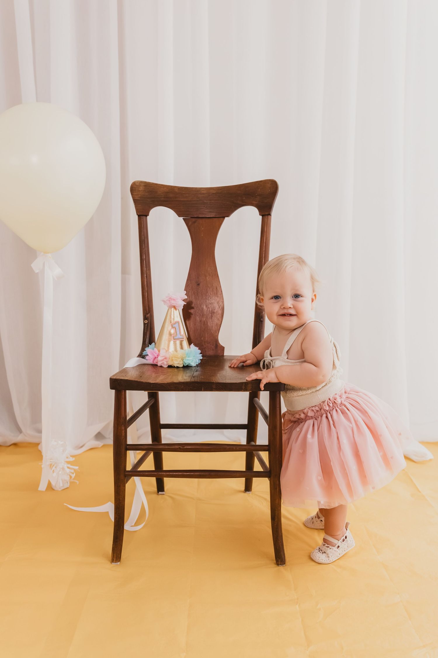 Baby girl in pink tutu dress stands by wooden chair with birthday cake topper and white balloon backdrop.