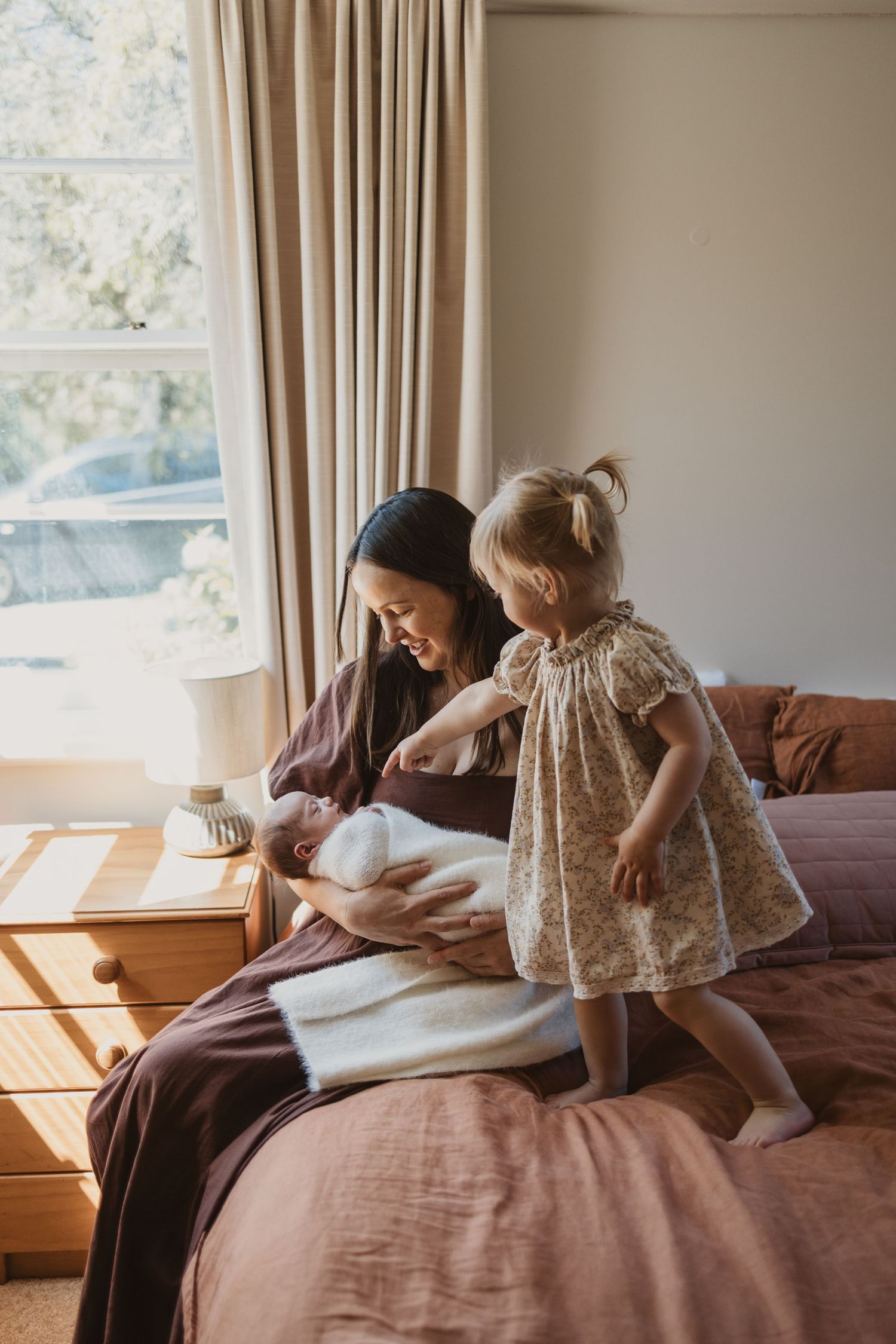 A mother sits on a bed holding a newborn while a toddler girl in a floral dress stands beside them in a sunlit bedroom.