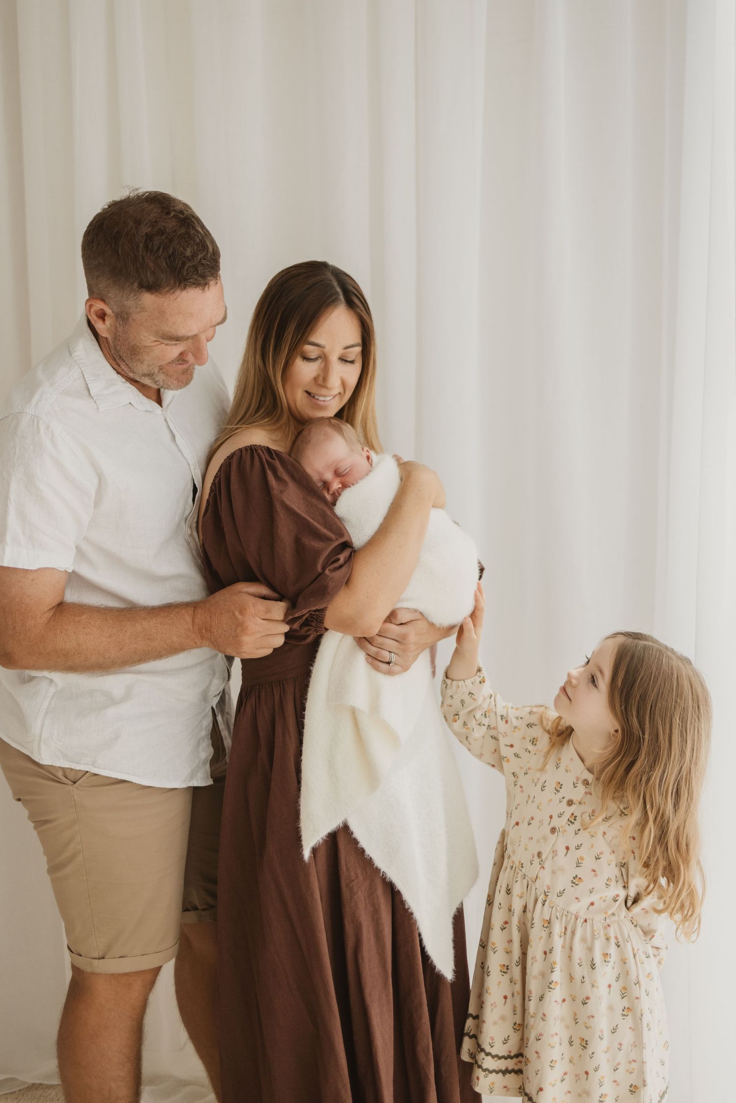 A family of four poses together, parents holding a newborn baby while their young daughter reaches up lovingly.