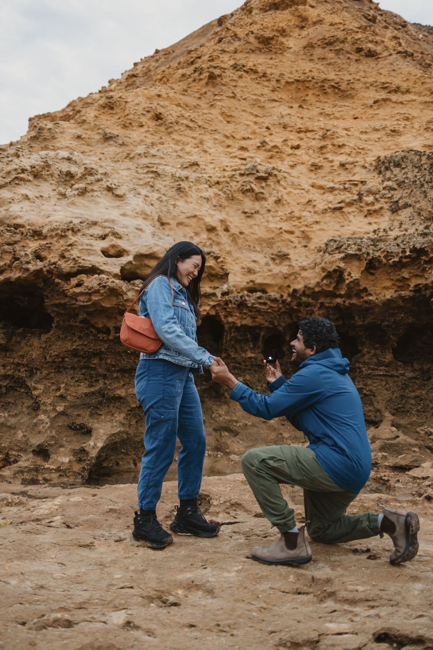 Man kneeling on one knee proposing to woman near sandy rock formation outdoors.