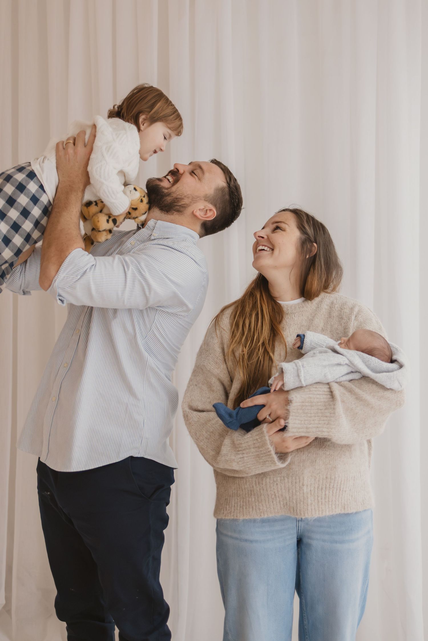 Happy family with father lifting toddler and mother holding newborn baby, smiling together against white curtain backdrop.