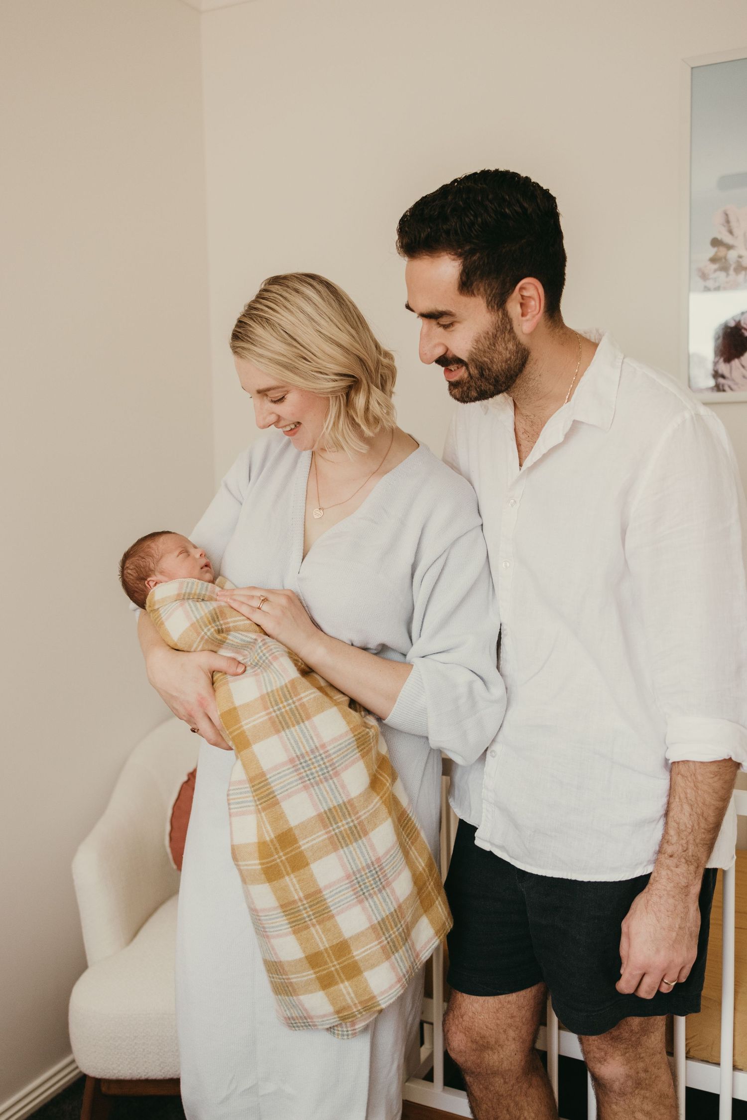 Parents in white clothing hold their newborn wrapped in yellow plaid blanket in nursery.