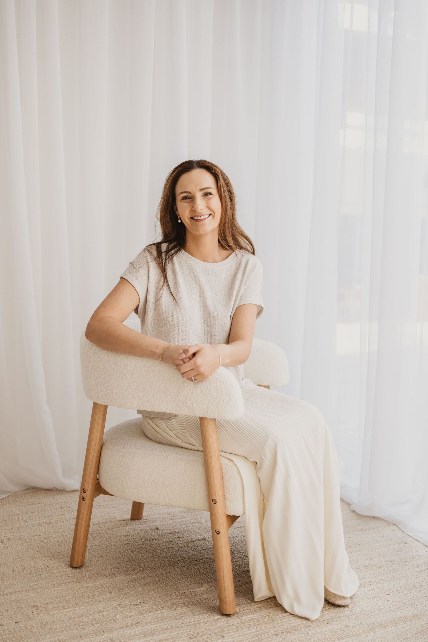 Smiling woman in beige outfit sitting relaxed on a cream armchair against white curtains in a bright room.