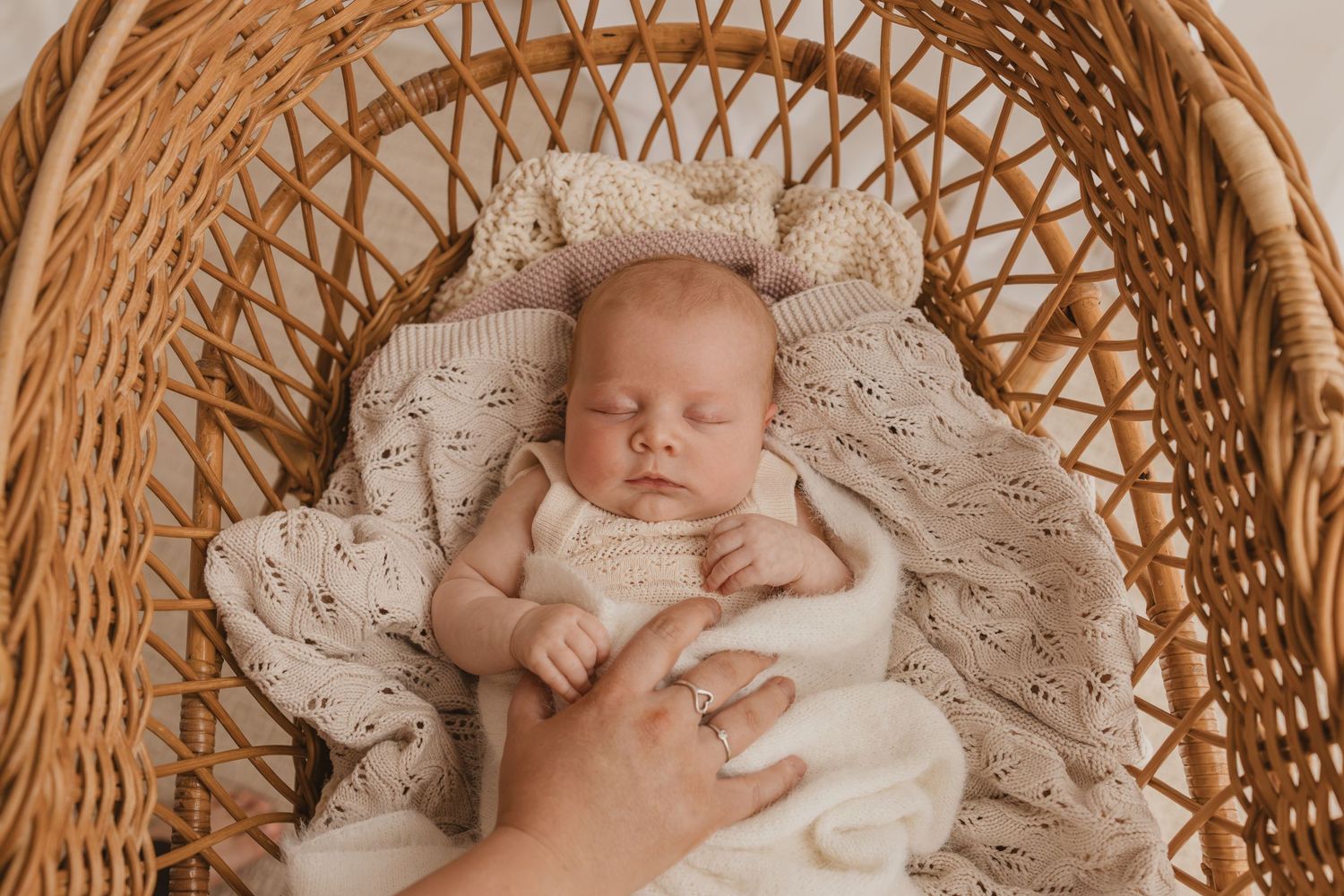 Newborn baby sleeping peacefully in a wicker bassinet, wrapped in a cream knit blanket, with a gentle hand resting nearby.