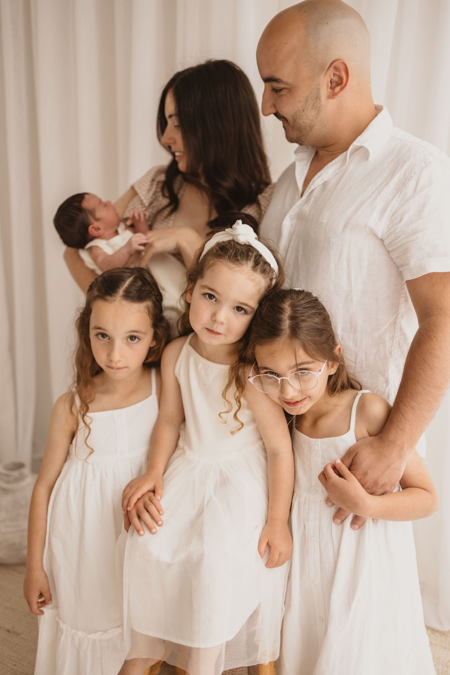 Family portrait with parents in white holding newborn, three young daughters in white dresses posing together indoors.