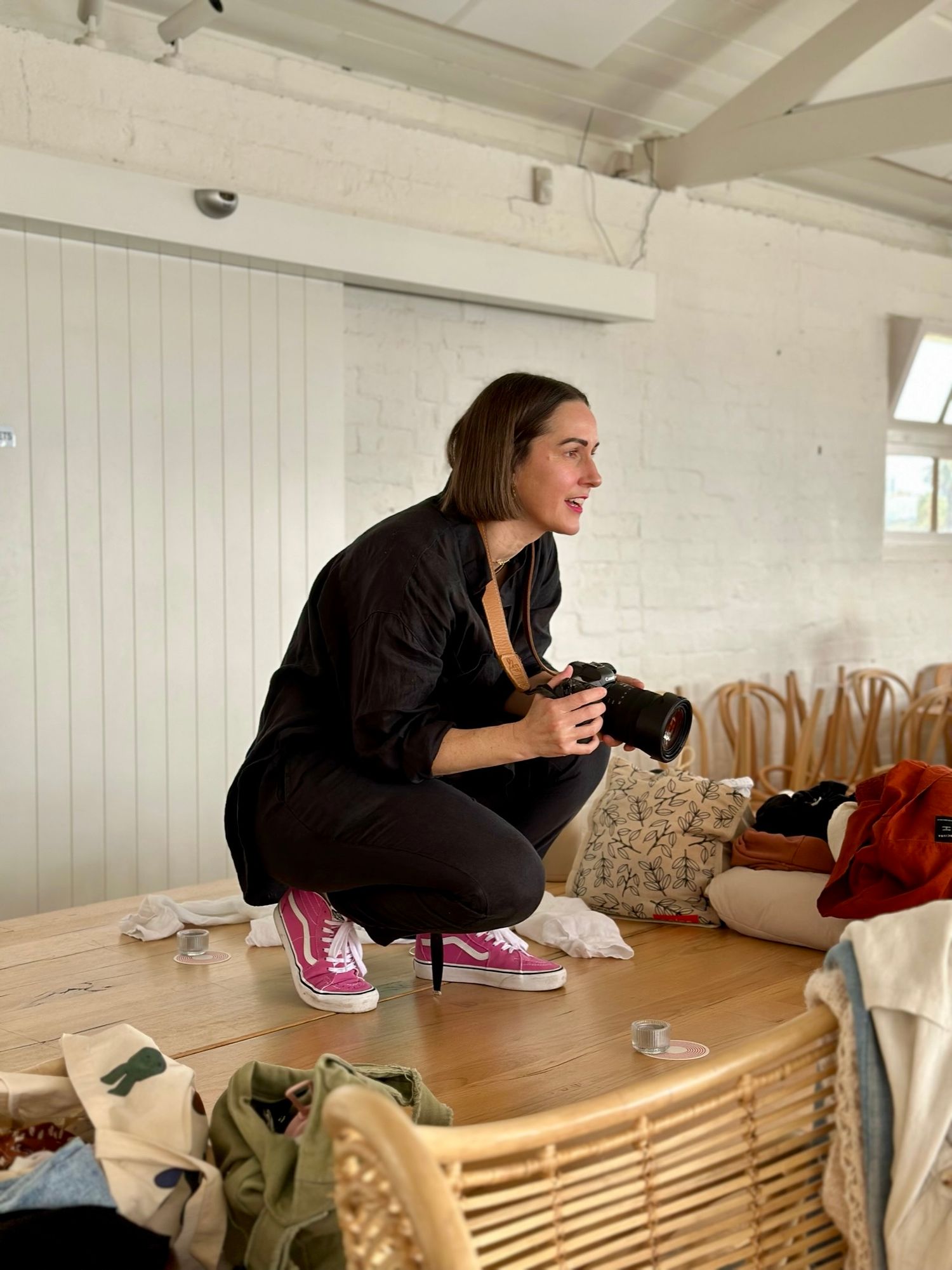 Woman in black outfit and pink Vans sneakers crouching with camera in bright white studio space.