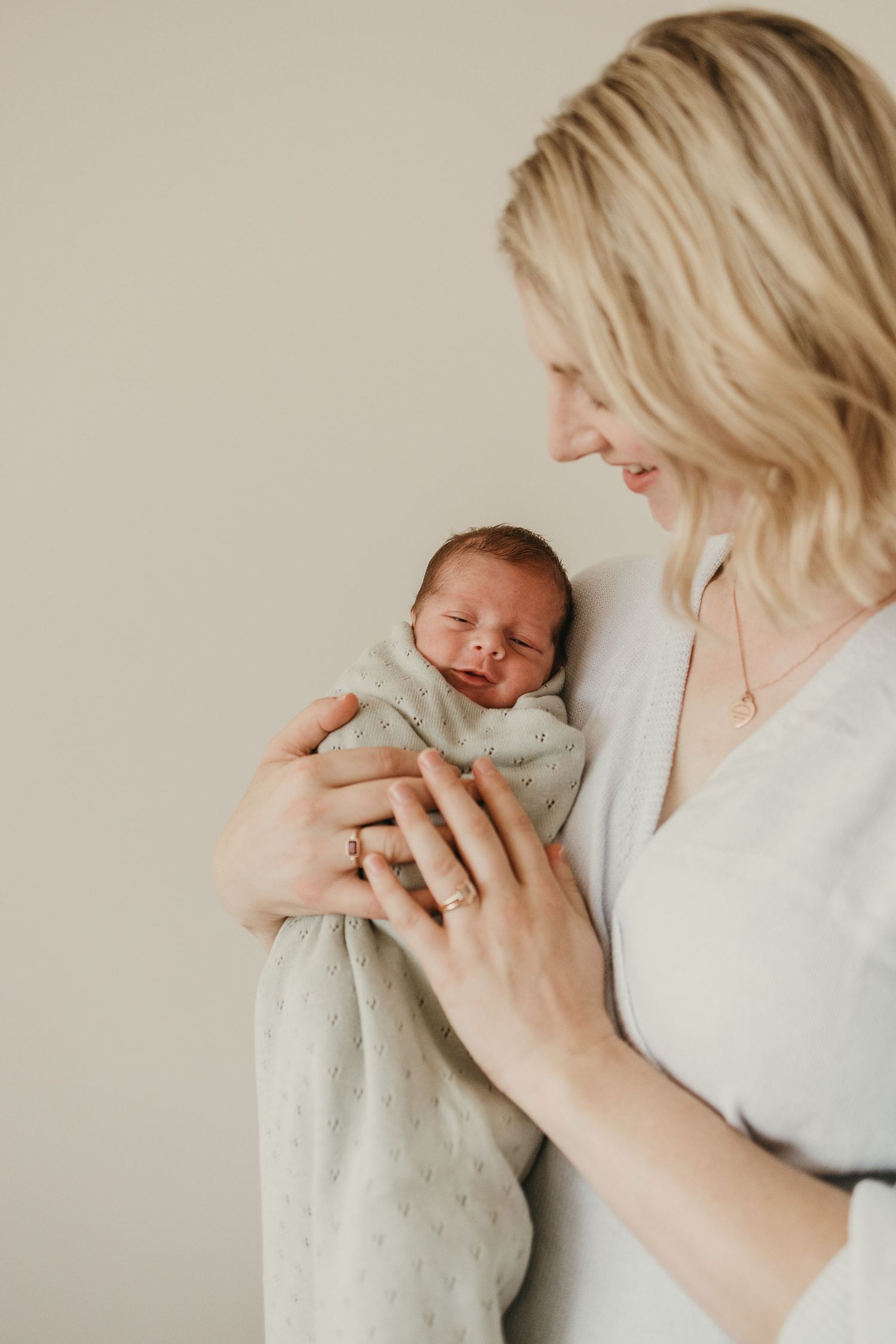 A tender moment captured between a parent and newborn baby swaddled in a light gray blanket against a neutral background.
