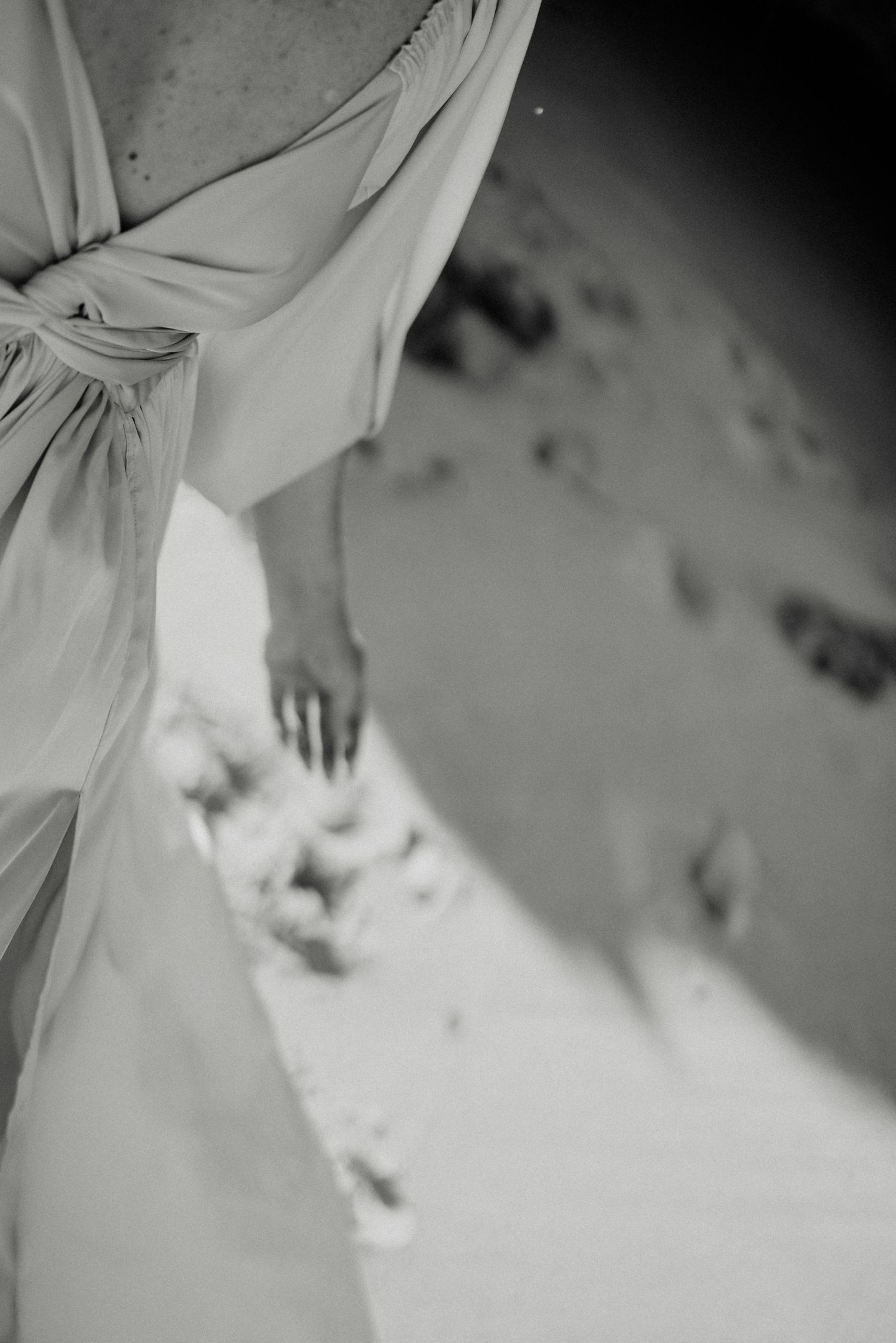 Black and white photo of a bride in a flowing wedding dress touching sandy beach ground with her hand.