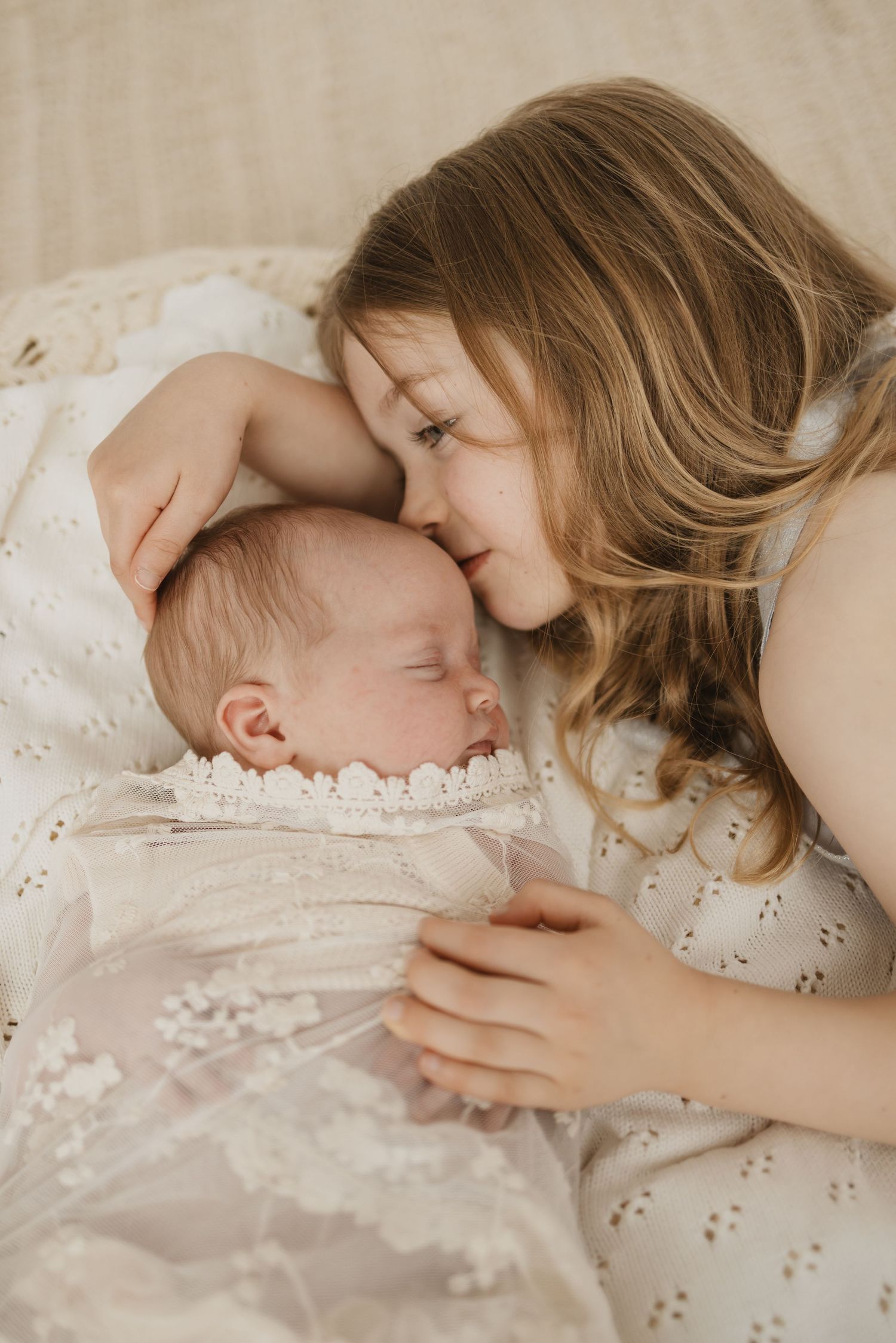 A young girl tenderly nuzzles a sleeping newborn baby wrapped in a delicate white lace swaddle.