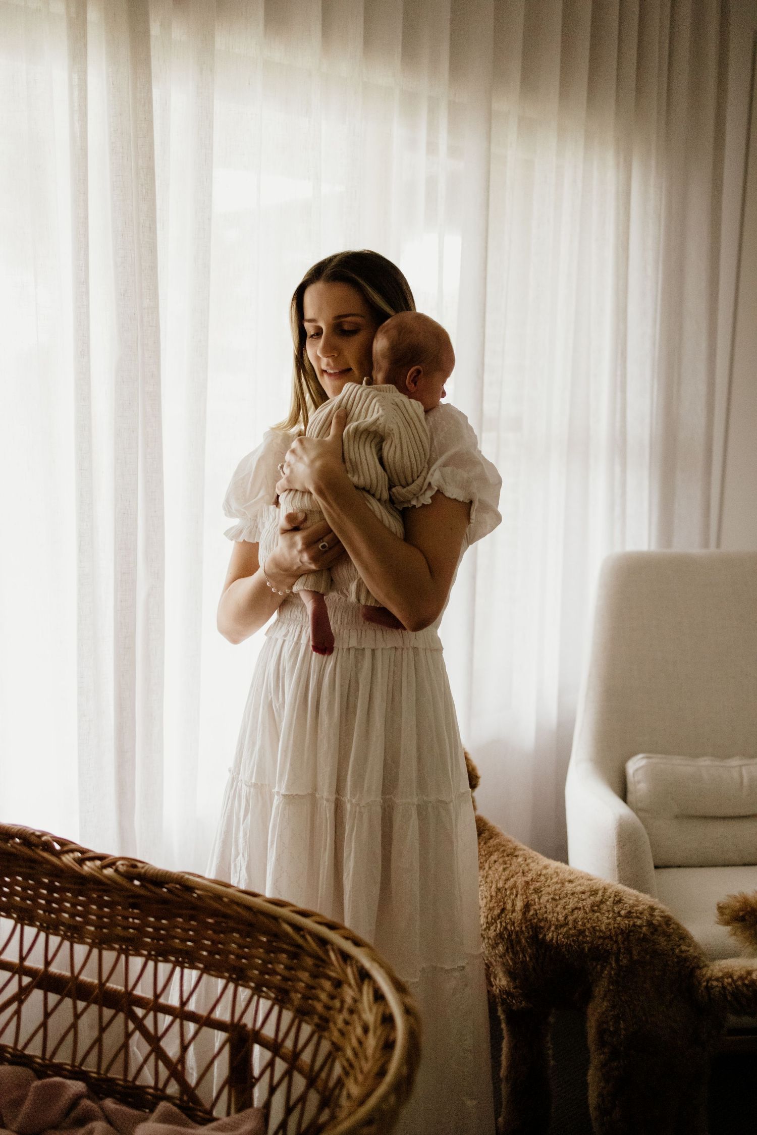 A mother in a white dress tenderly holds her newborn baby near a sunlit window in a soft, neutral nursery.