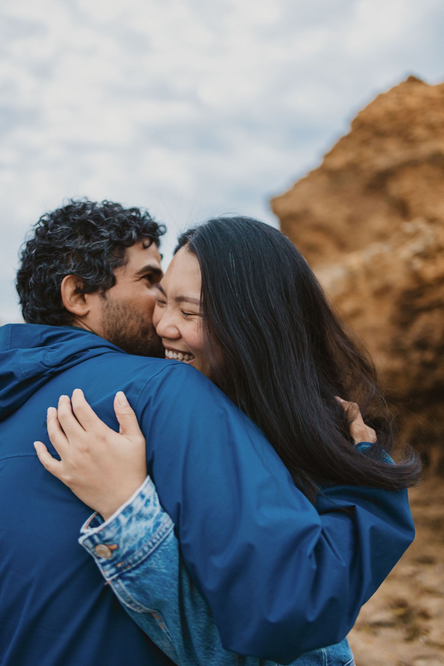 A joyful couple embracing and laughing outdoors near rocky cliffs, both wearing blue jackets.