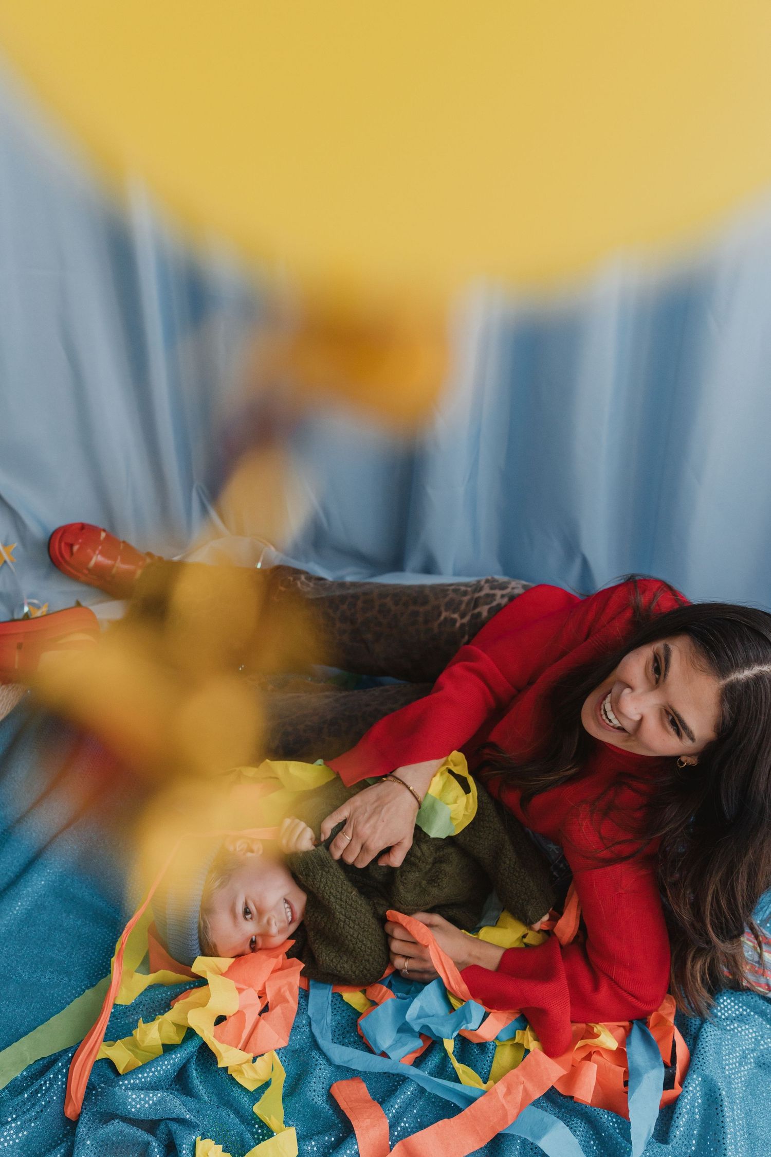 A smiling woman in red lies on a blue blanket with a baby surrounded by colorful ribbons and a yellow backdrop.