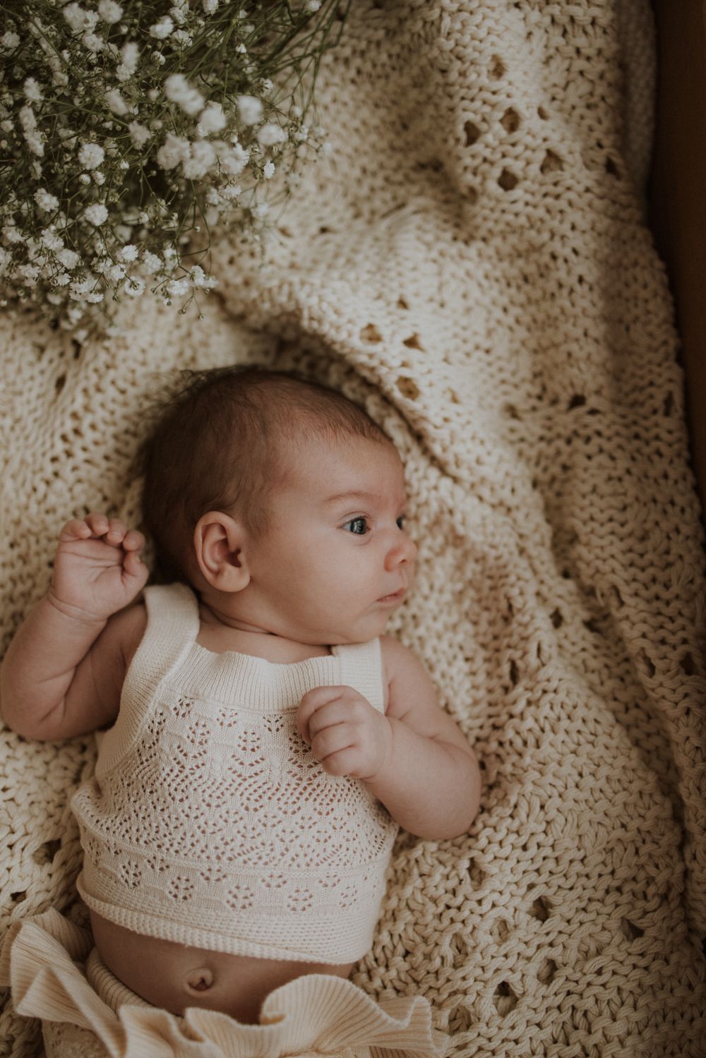 Newborn baby lying on a cream knit blanket wearing a white lace romper, with delicate white flowers nearby.