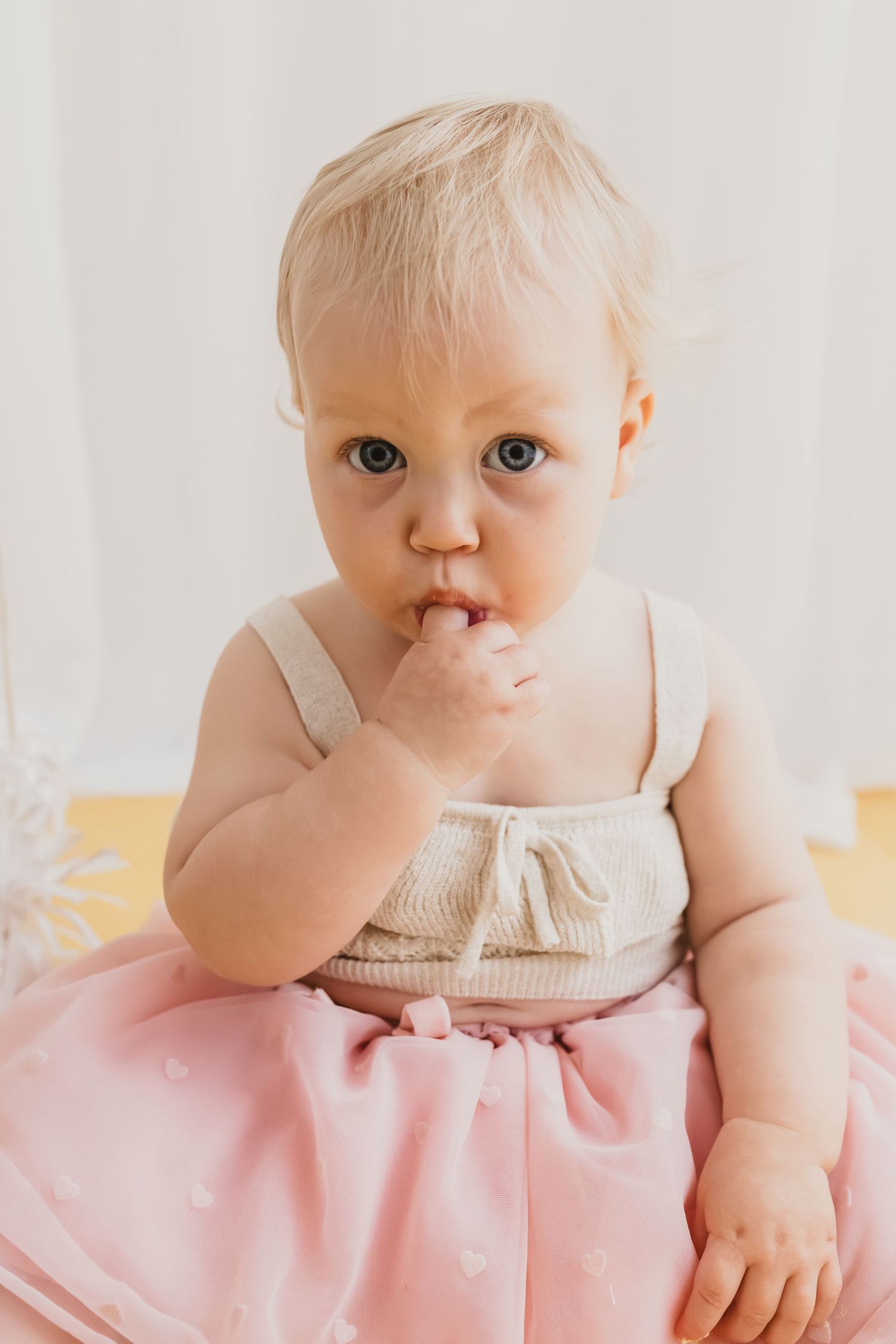Adorable blonde baby girl in pink tutu dress and white top sitting and putting fingers in mouth.