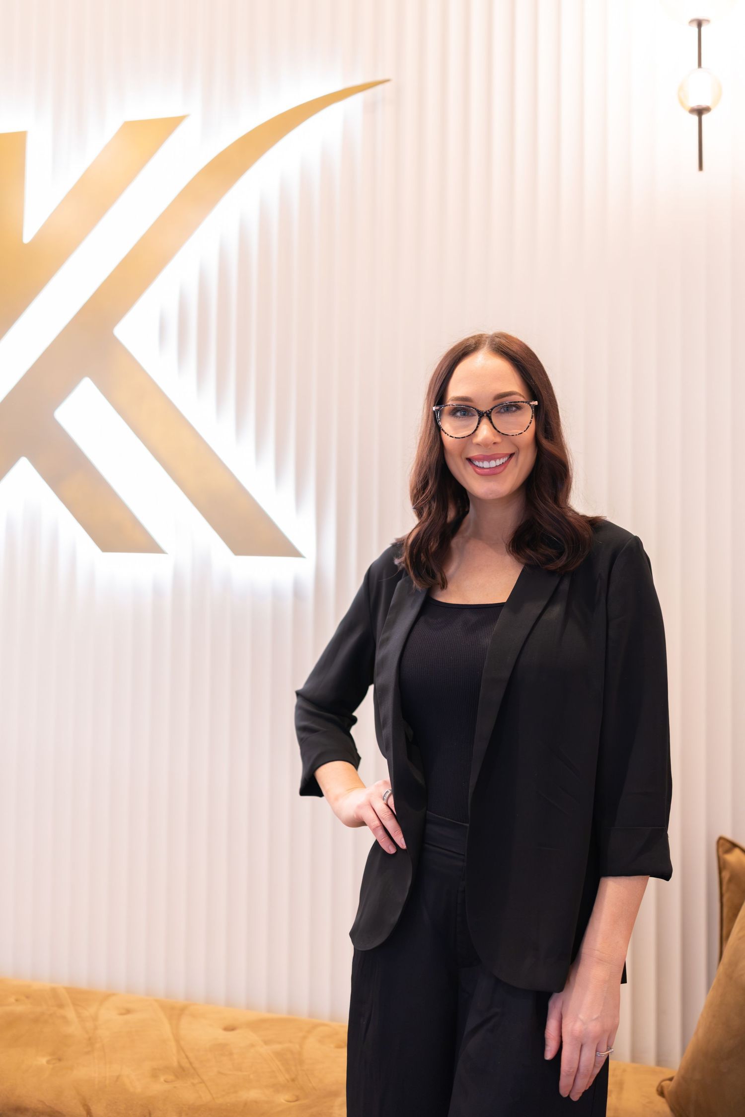 A smiling woman in a black blazer and glasses stands confidently in front of a gold K logo on a white wall.