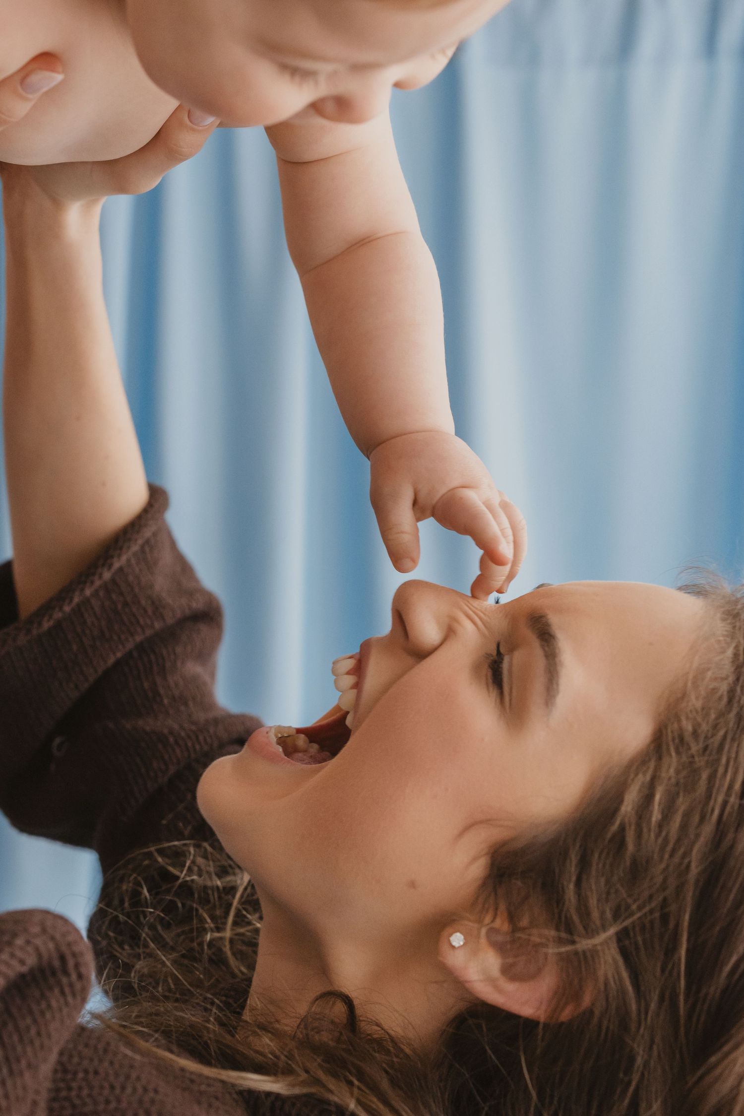 A joyful mother lifts her smiling baby overhead, sharing a tender moment against a soft blue background.