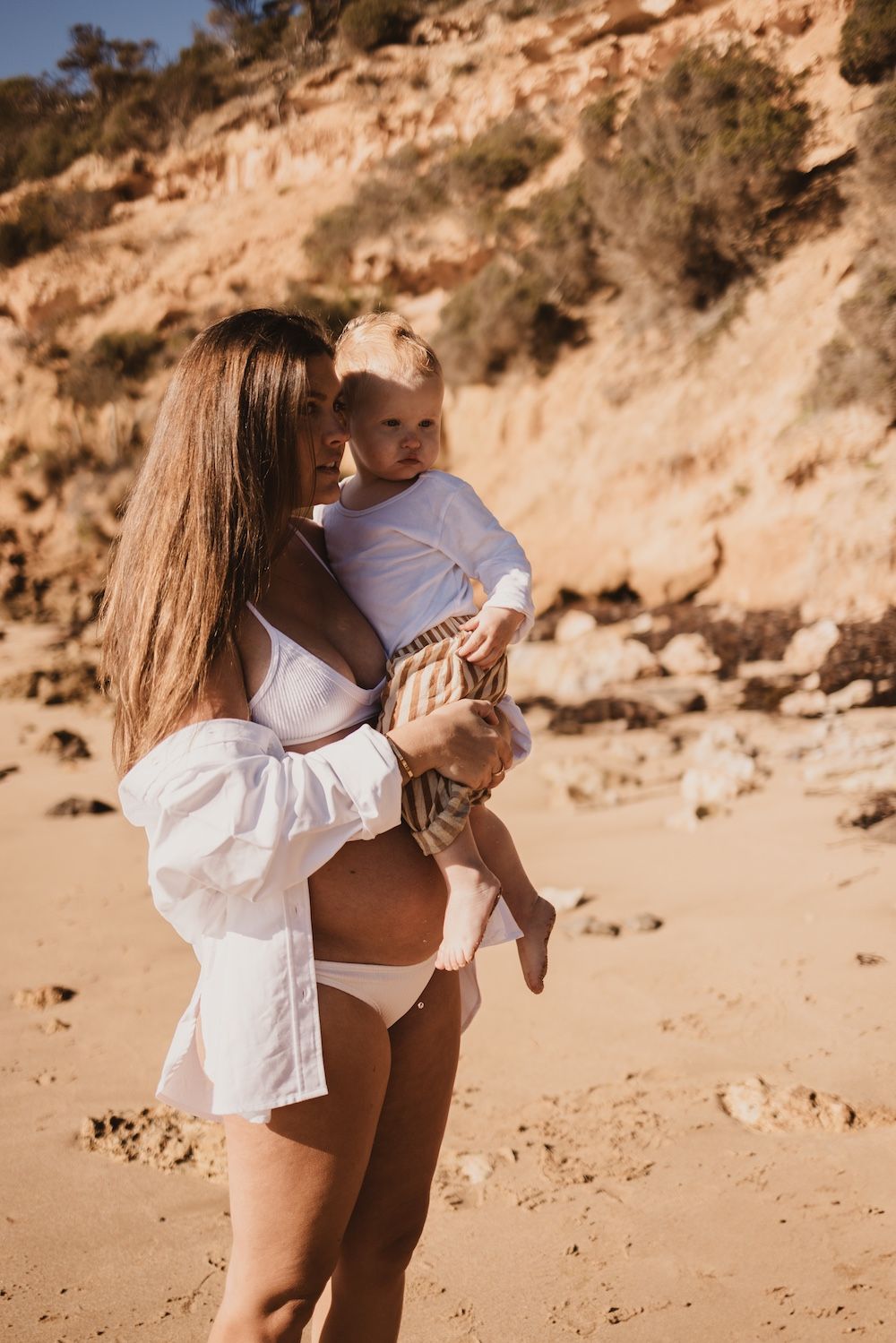 A mother in a white bikini and shirt holds her baby on a sandy beach with rocky cliffs in the background.