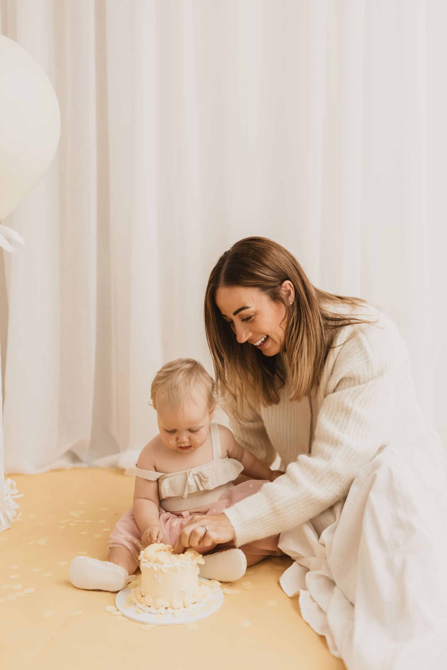 Smiling mother helping her baby girl taste a white smash cake during first birthday celebration.