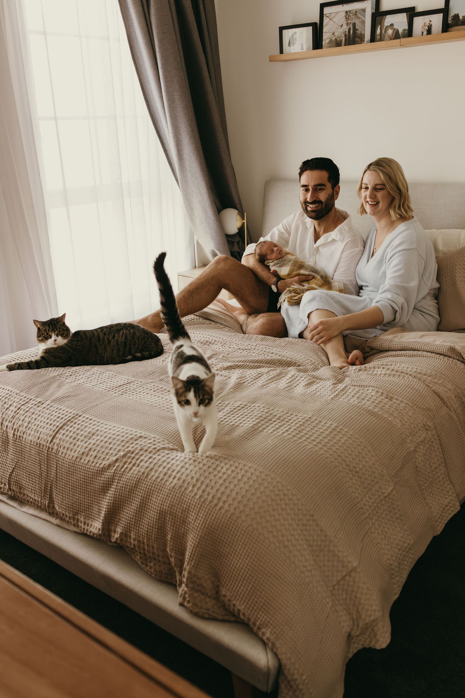 Family relaxing on bed with two cats while holding their newborn baby.