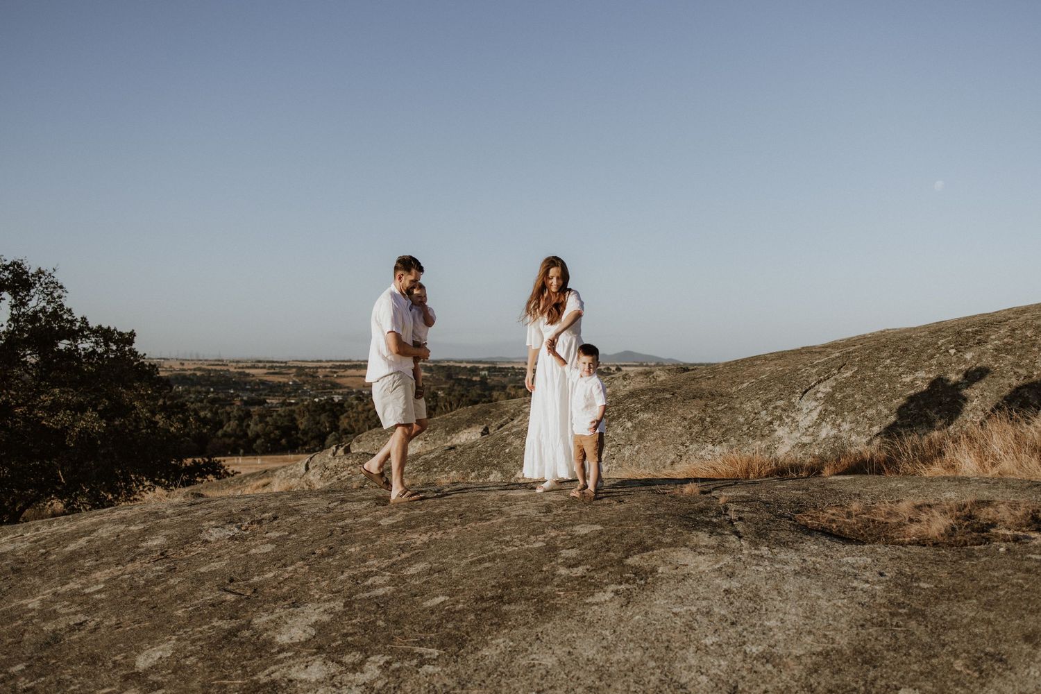 Couple walking barefoot on rocky hilltop overlooking scenic valley landscape.