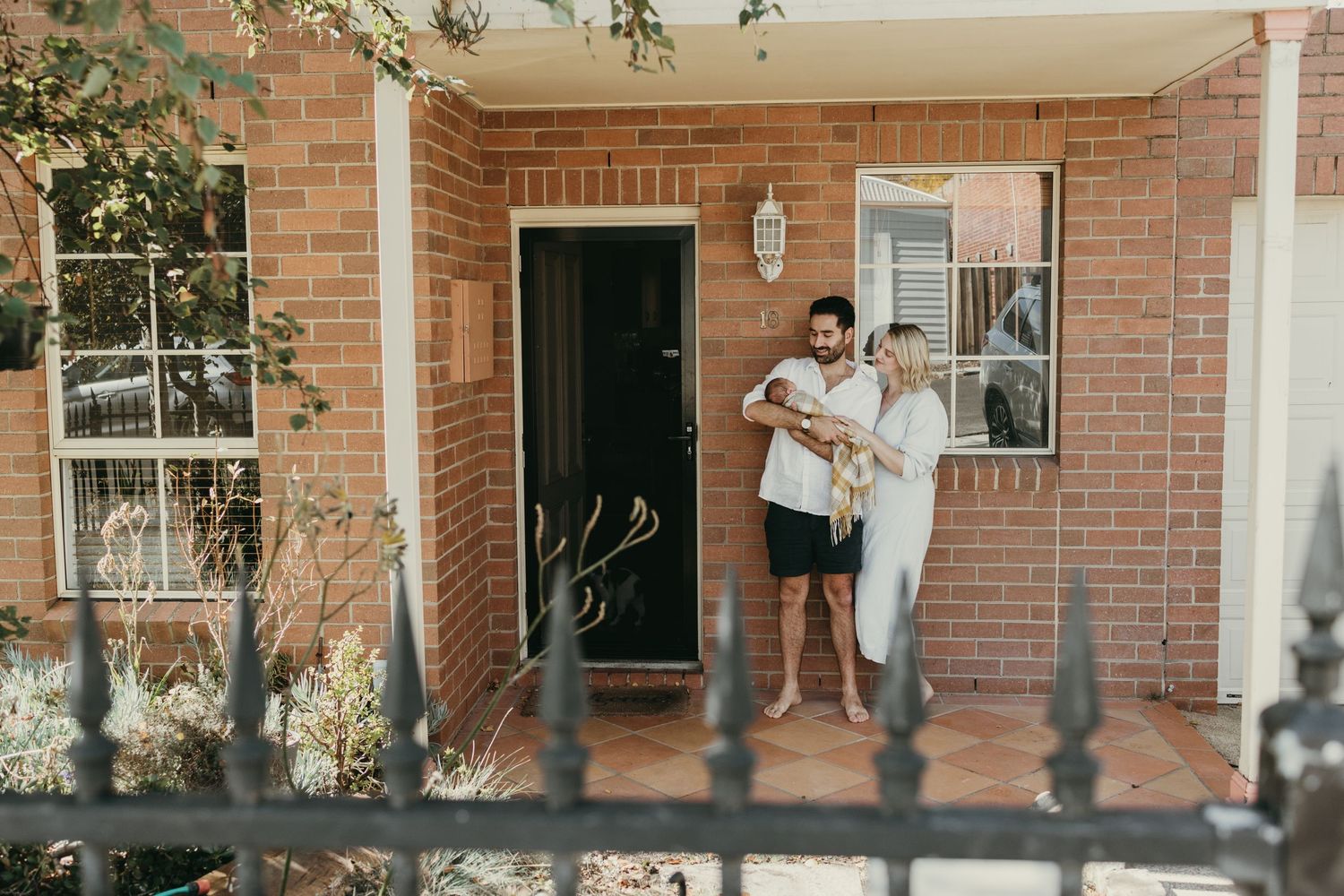 Couple standing on front porch of brick house while holding their newborn.