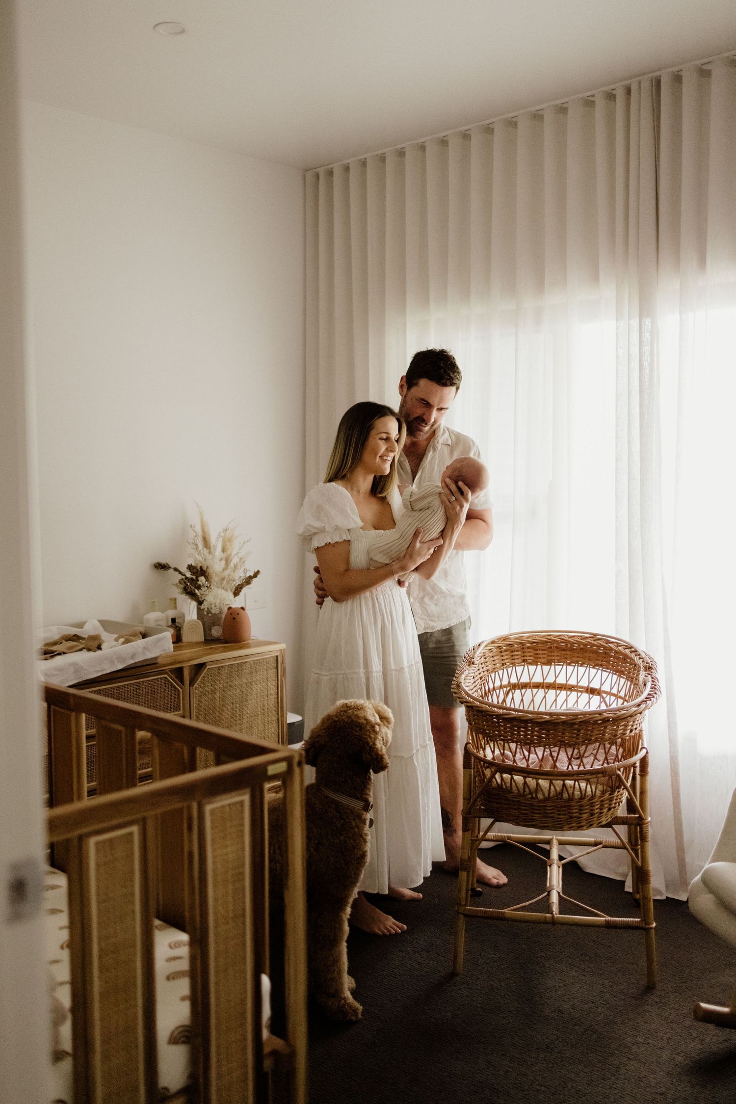 A couple in a boho nursery admiring their newborn baby, with a dog nearby and a rattan crib and chair.