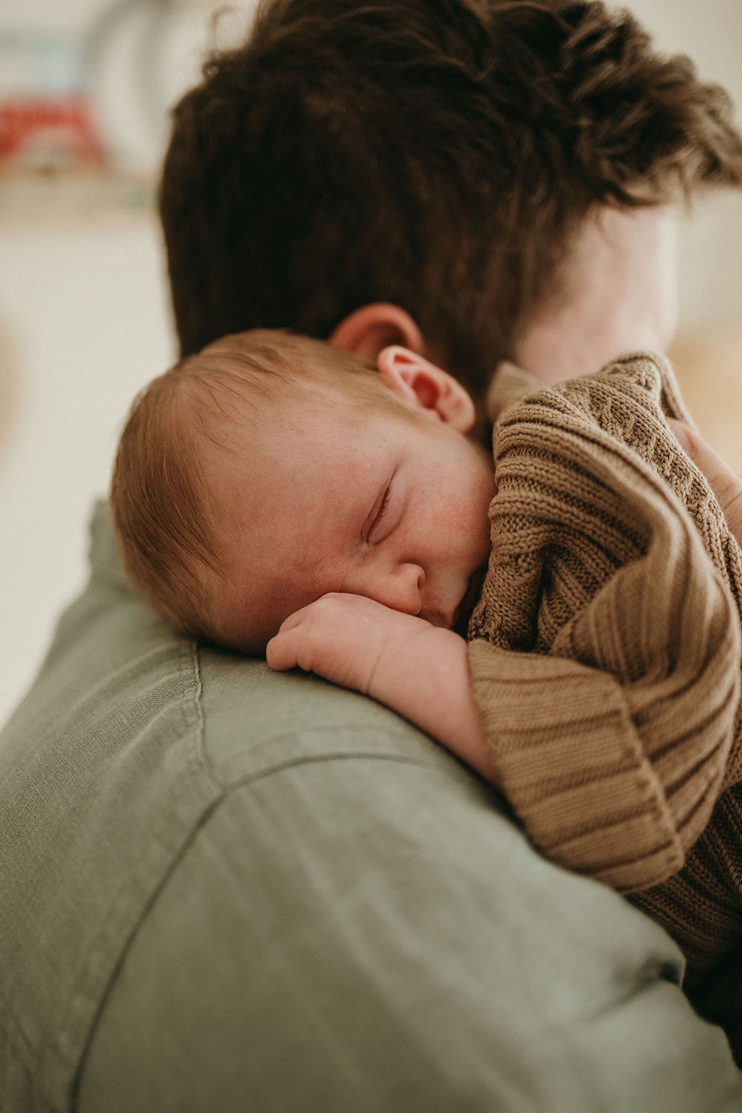 A newborn baby sleeping peacefully on a parent's shoulder, wrapped in a cozy brown knit outfit.