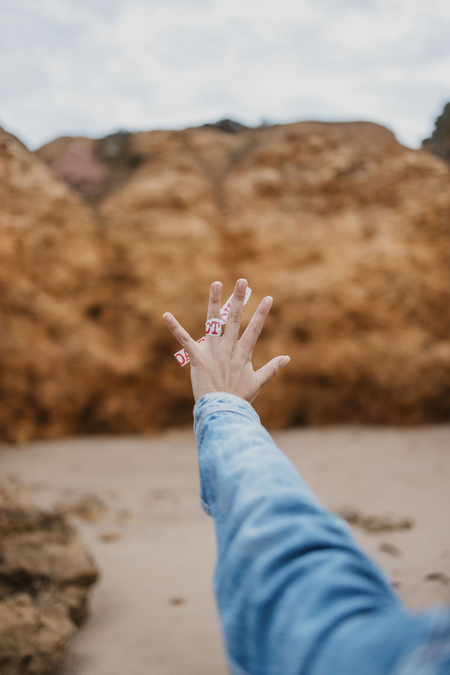 A hand with pink rings reaches out toward rocky cliffs on a sandy beach, wearing a blue denim jacket sleeve.