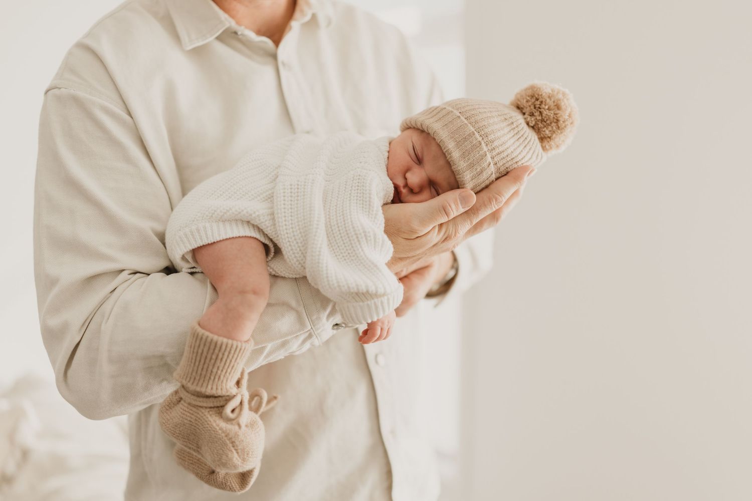 Adult cradling a sleeping newborn baby dressed in a white knit outfit and beige pom-pom hat.