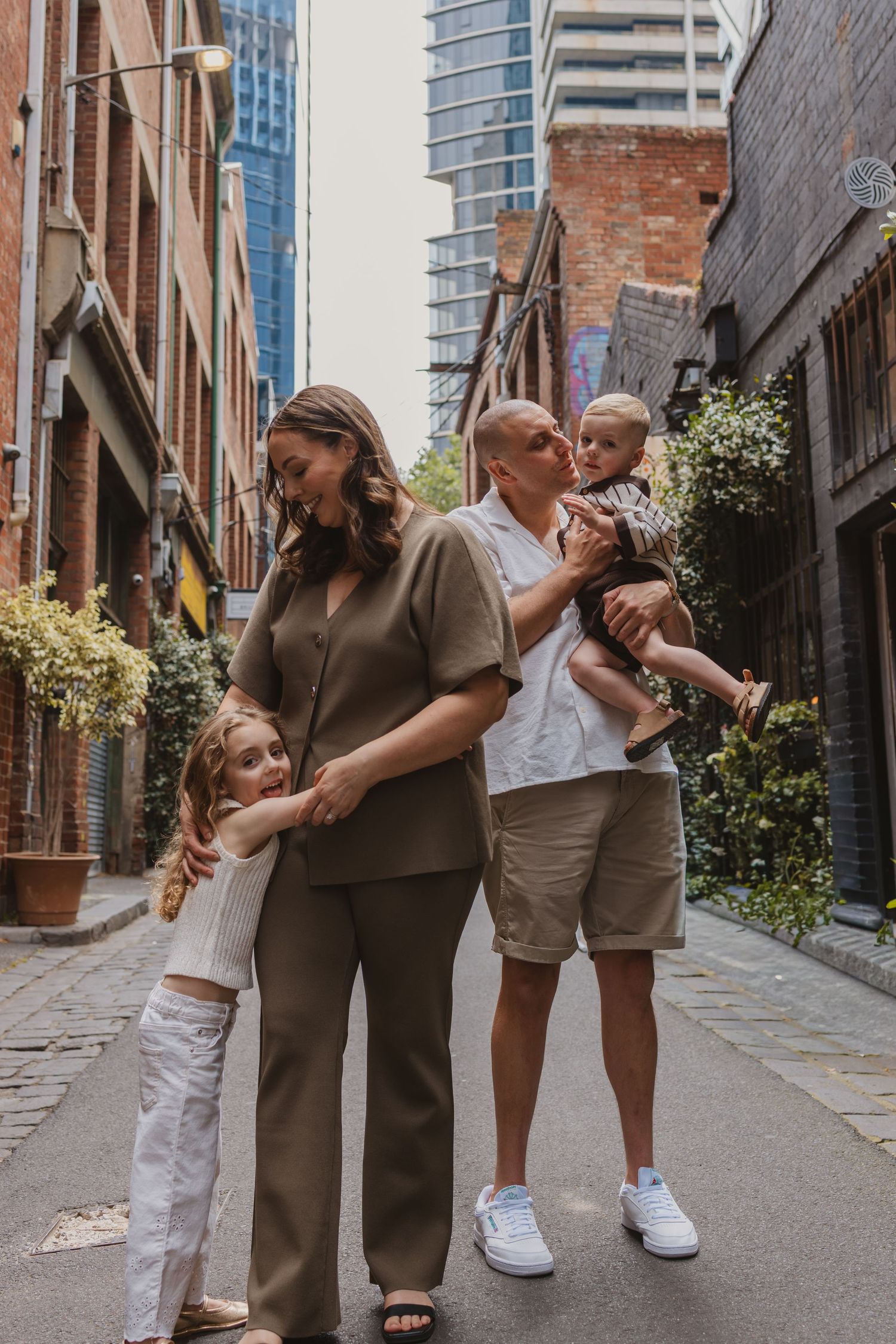 Family of four posing in a urban laneway, parents holding children, surrounded by brick buildings and skyscrapers.