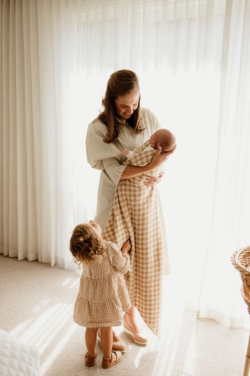 A mother in a checkered dress holds a newborn while her toddler daughter reaches up to her in a bright room.