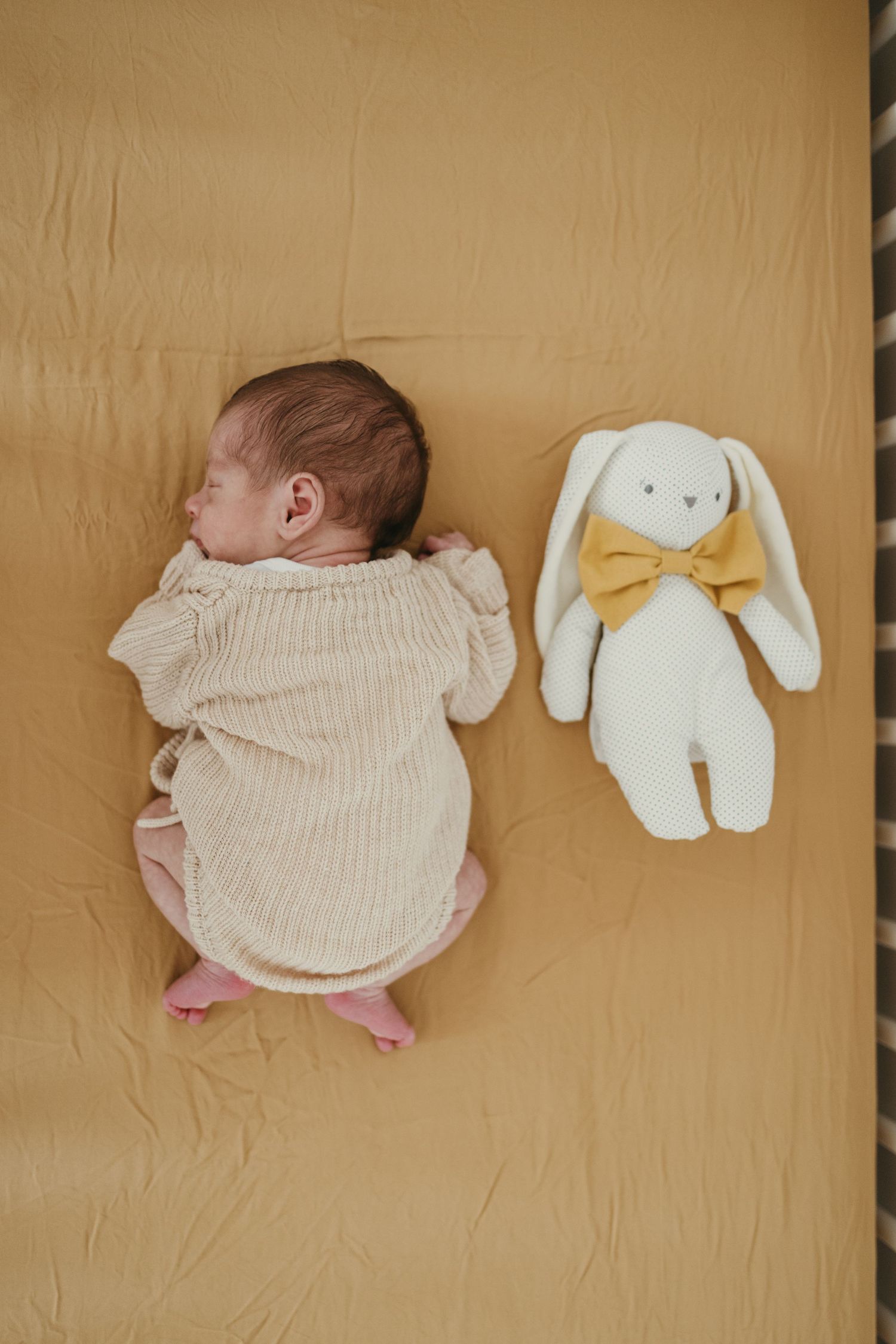 Sleeping newborn in knit sweater next to white plush bunny toy on beige background.