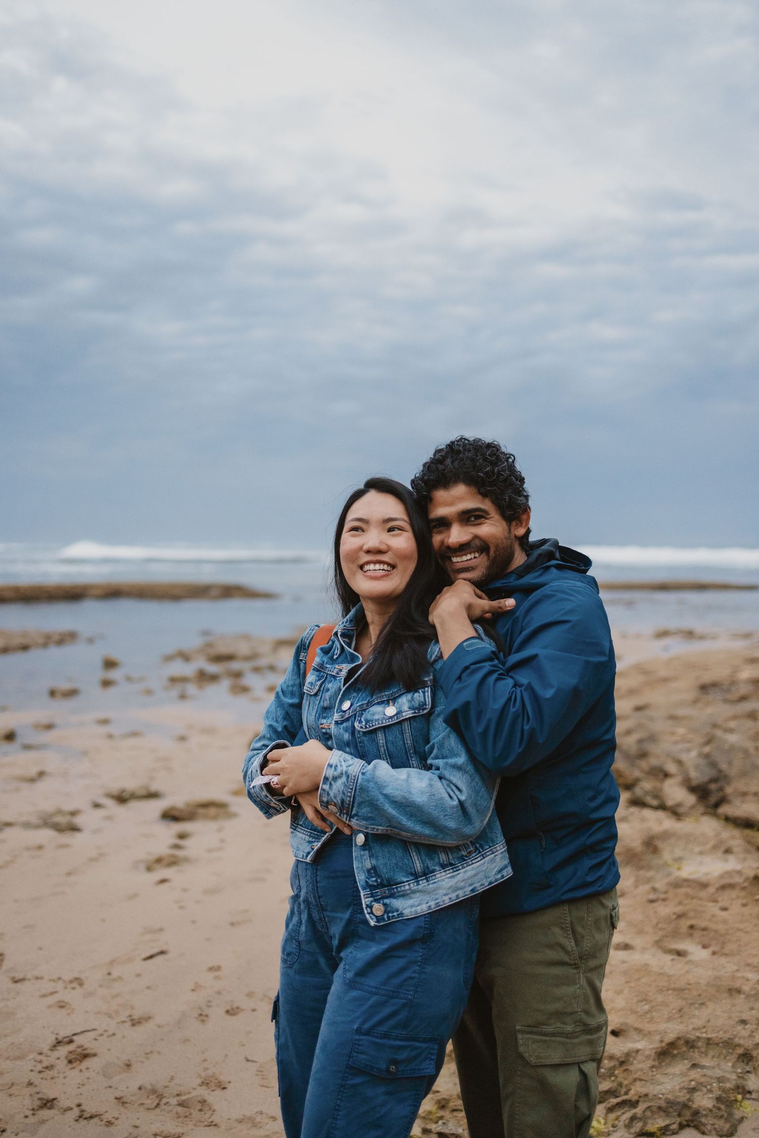Happy couple embracing on a sandy beach, woman in denim outfit and man in blue jacket, with ocean waves behind them.