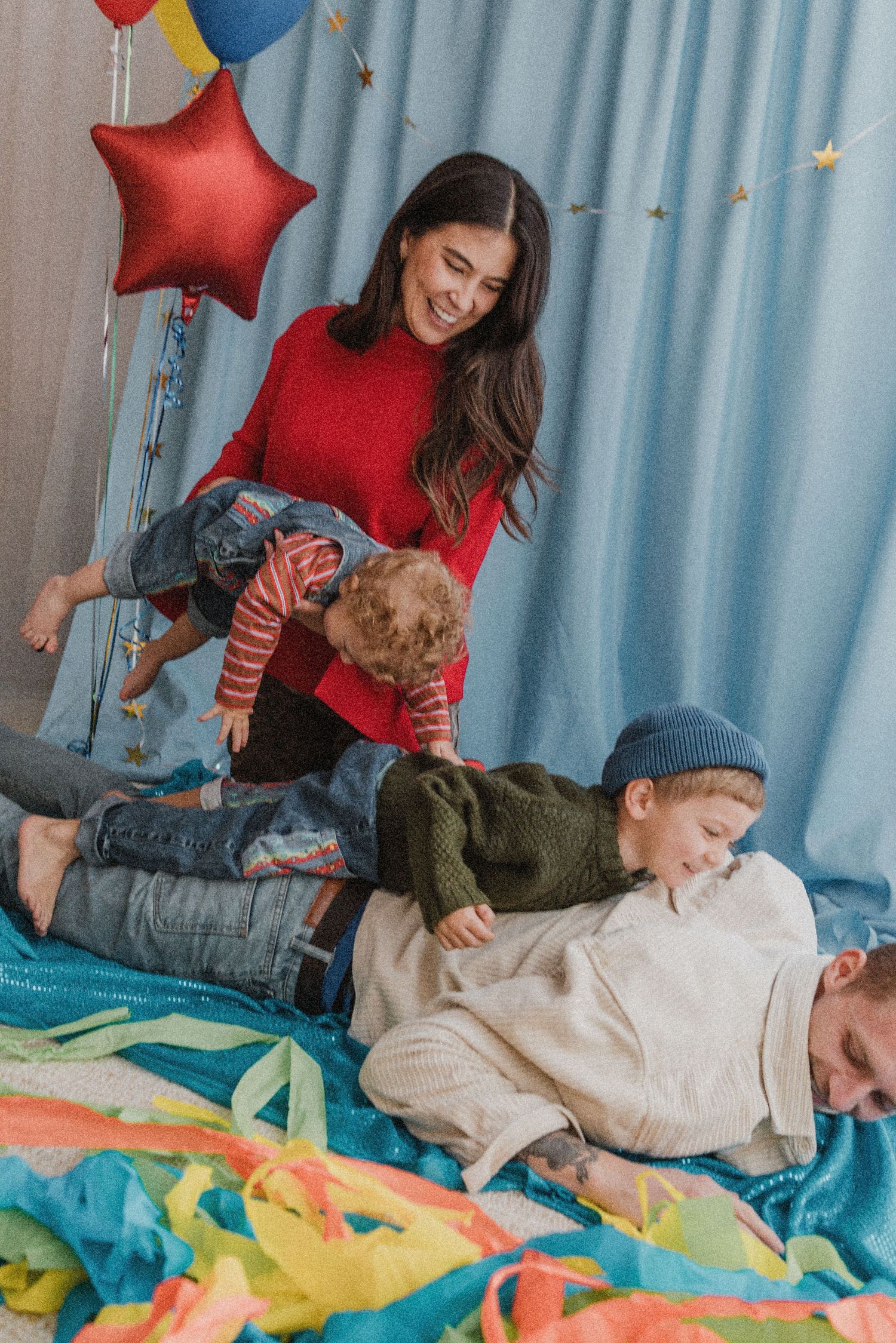 A smiling woman in a red sweater plays with two young children on a colorful blanket surrounded by festive star balloons.