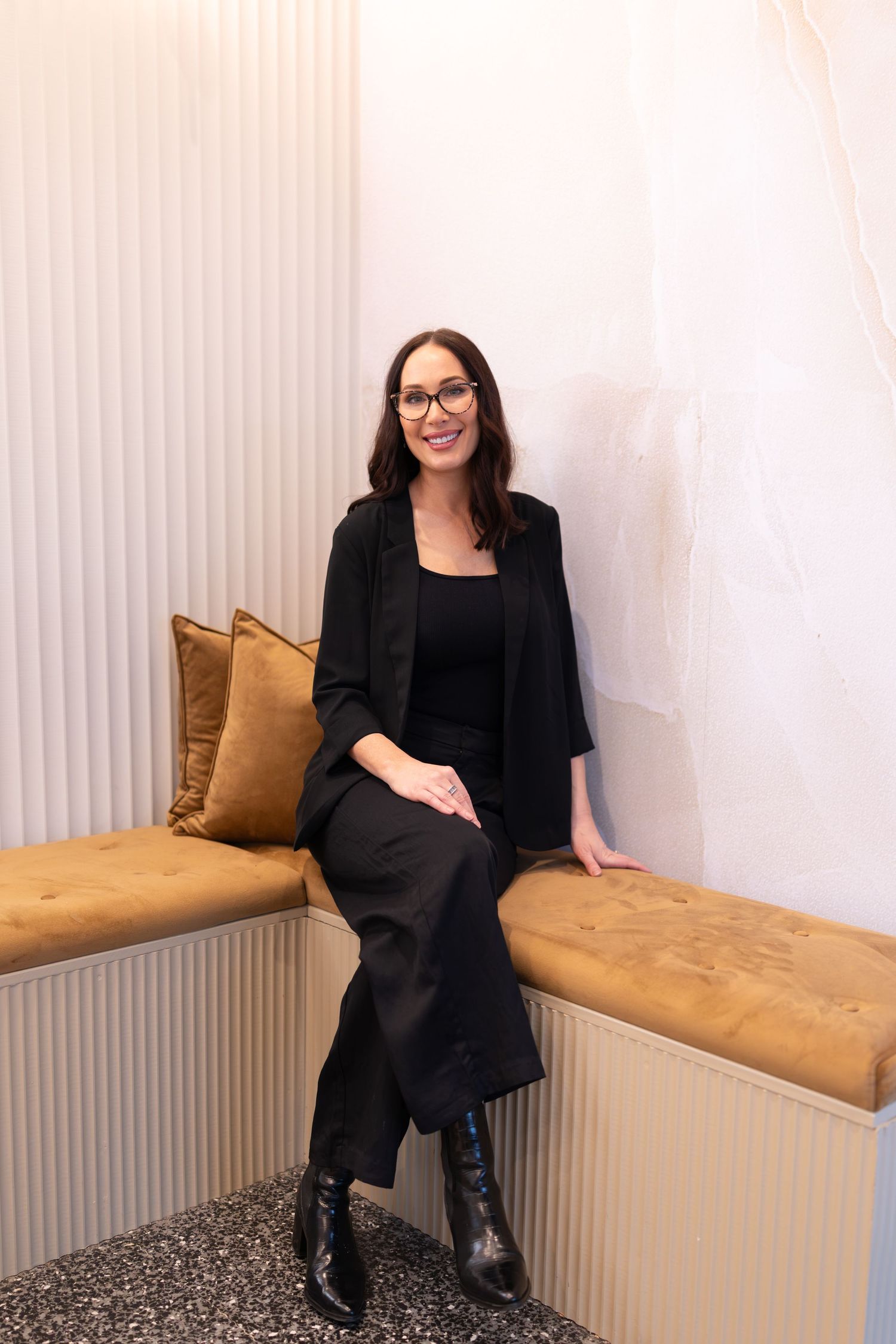 Woman in black blazer and pants sitting on a gold velvet bench, smiling, wearing glasses.