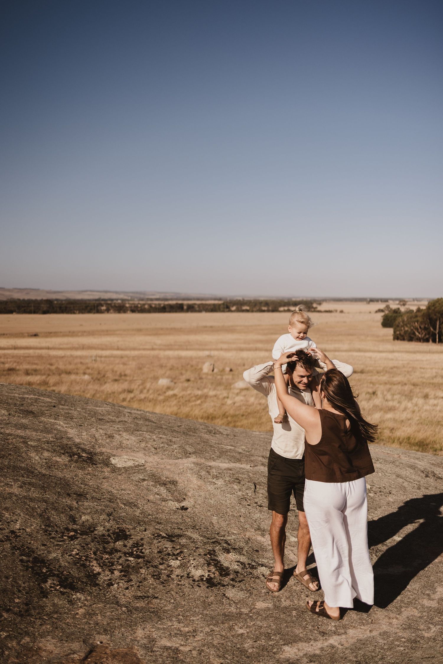 Family of three enjoying a sunny day in golden wheat fields, father carrying toddler on shoulders with mother beside them.