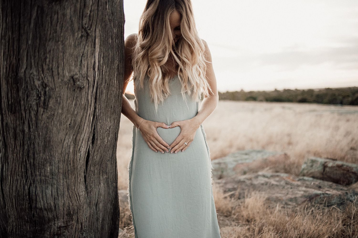 Pregnant woman in sage green dress forming heart shape on baby bump beside tree in golden field.