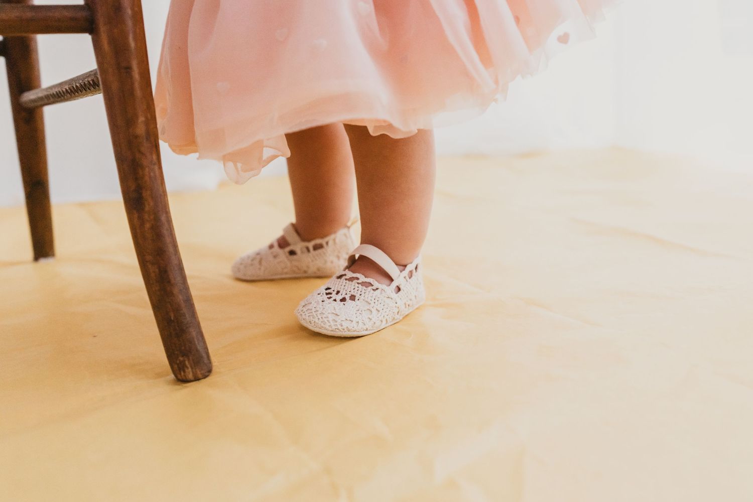 Close-up of a toddler in a pink tulle dress wearing white lace Mary Jane shoes on a yellow floor.