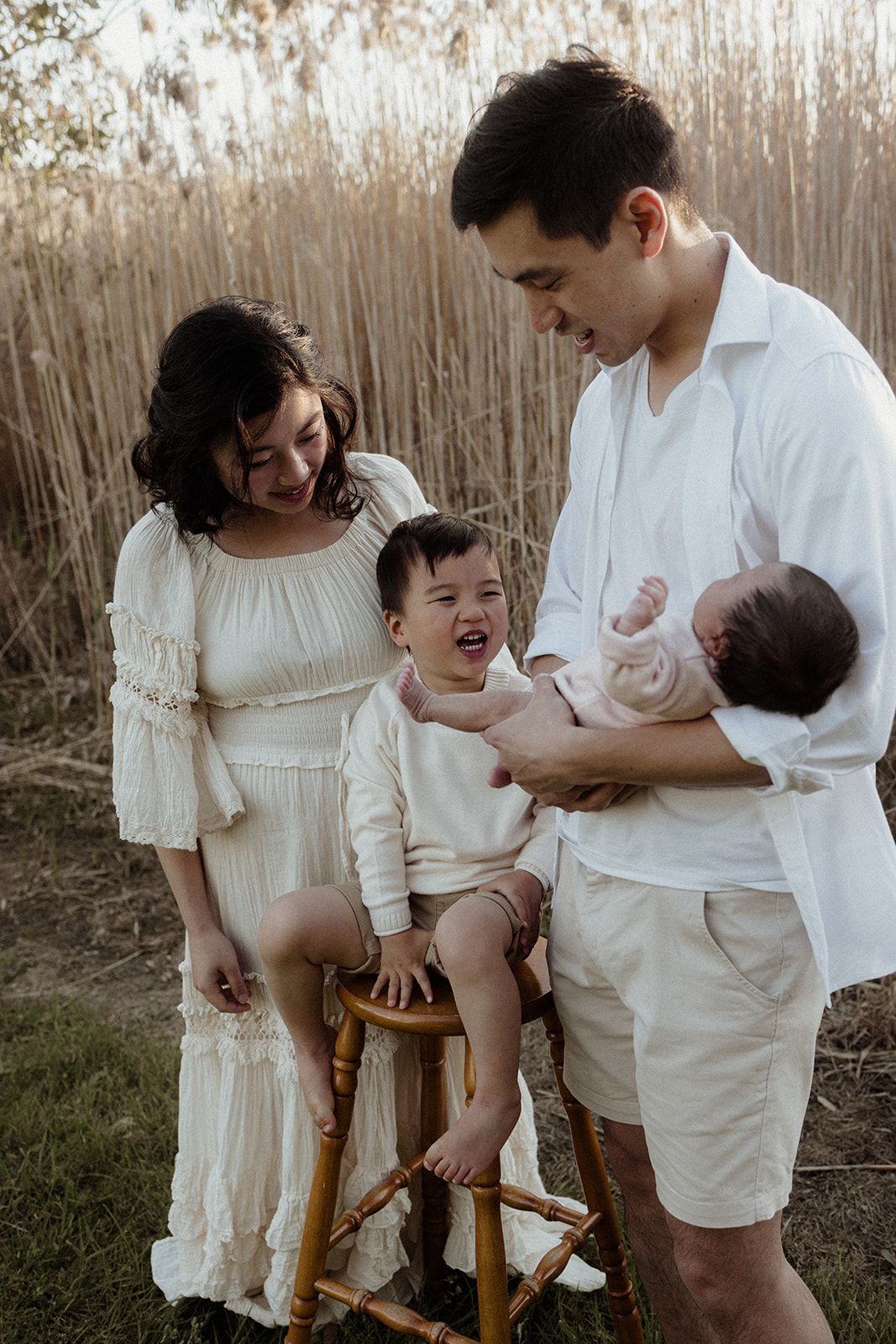 Family of four in white outfits posing outdoors near tall reeds, parents smiling at laughing toddler and newborn baby.